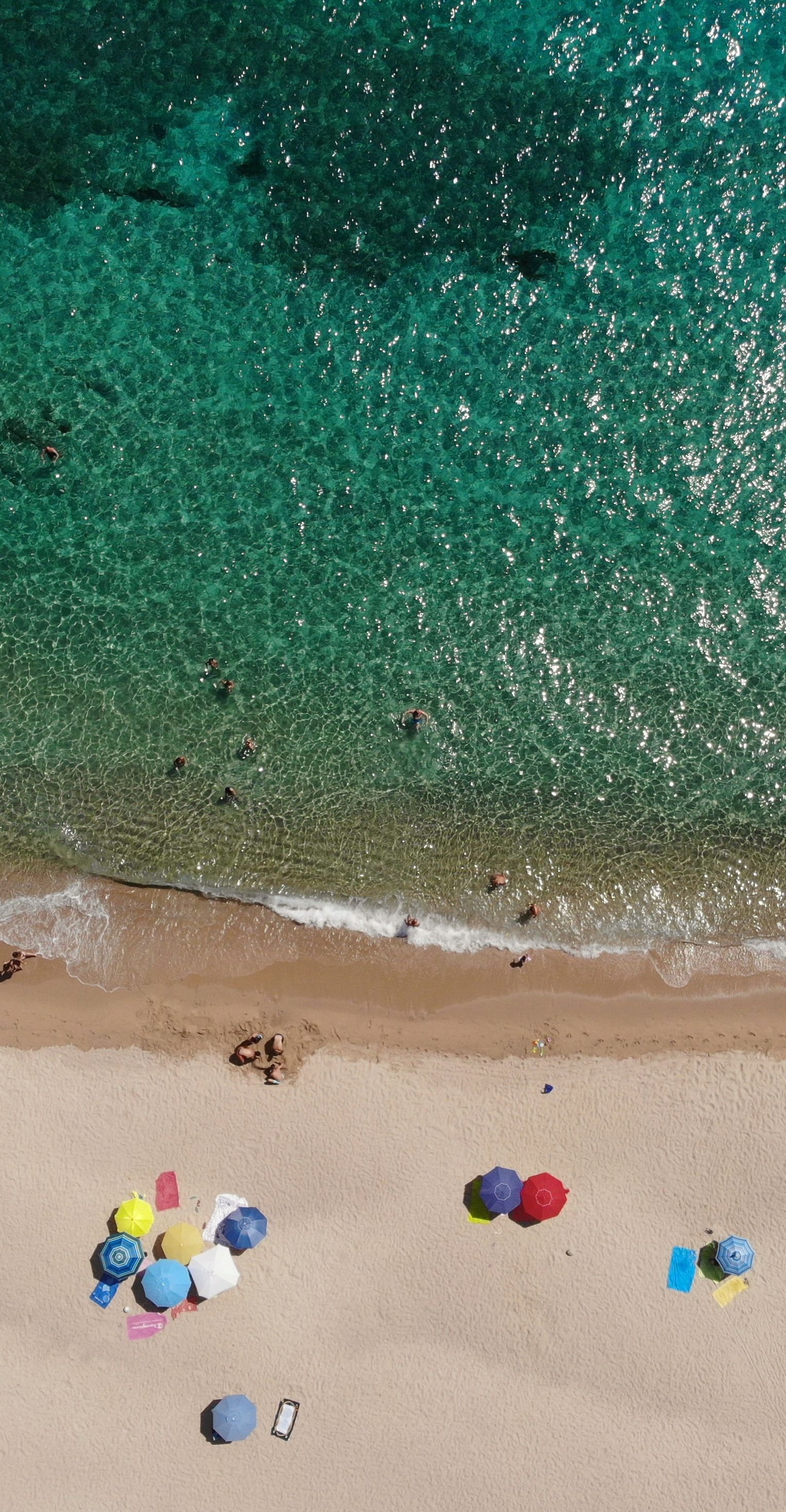 Aerial view of a sandy beach with colorful umbrellas and people swimming and relaxing near the shoreline.