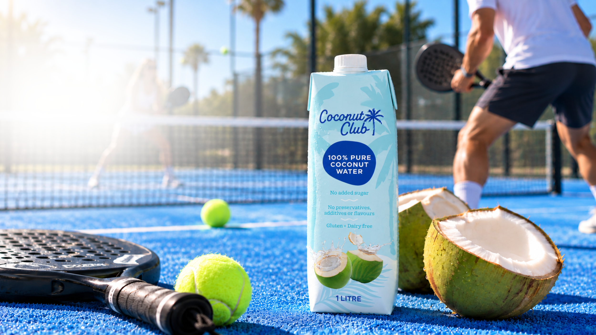 A carton of coconut water on a blue tennis court with tennis balls, a tennis racket, and a halved coconut in the foreground, while a person plays tennis in the background.