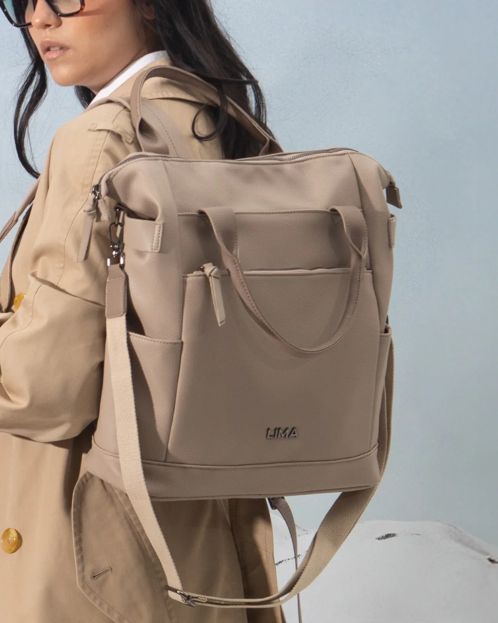 Mujer con gafas oscuras lleva un saco color beige y una mochila tipo tote beige con la marca 'LIMA' en la parte inferior, en un fondo de pared azul claro.
