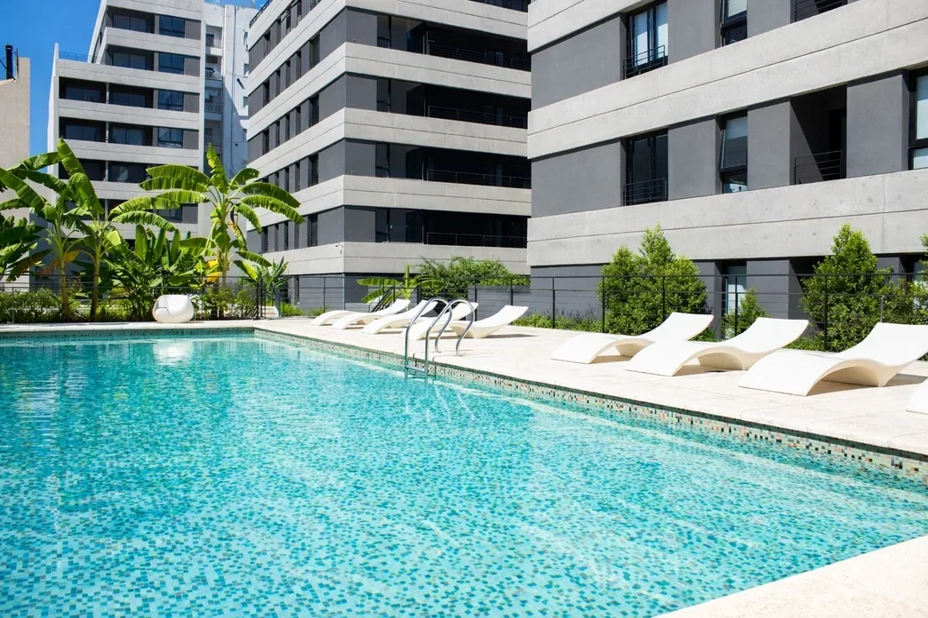 Piscina en un área residencial moderna con tumbonas blancas y plantas tropicales.