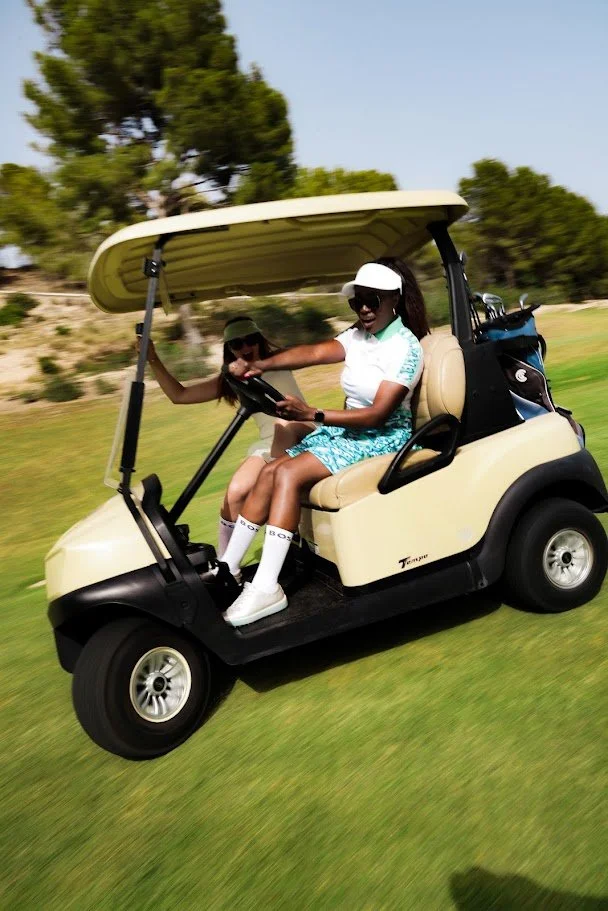 Dos jóvenes mujeres jugando golf en un carrito en un campo de golf, una de ellas sostiene un putter y parecen estar pasando un buen rato.
