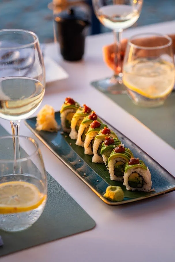 Plato de sushi con varias piezas enrolladas, acompañadas de jarras de agua con limón y vino en una mesa al aire libre.
