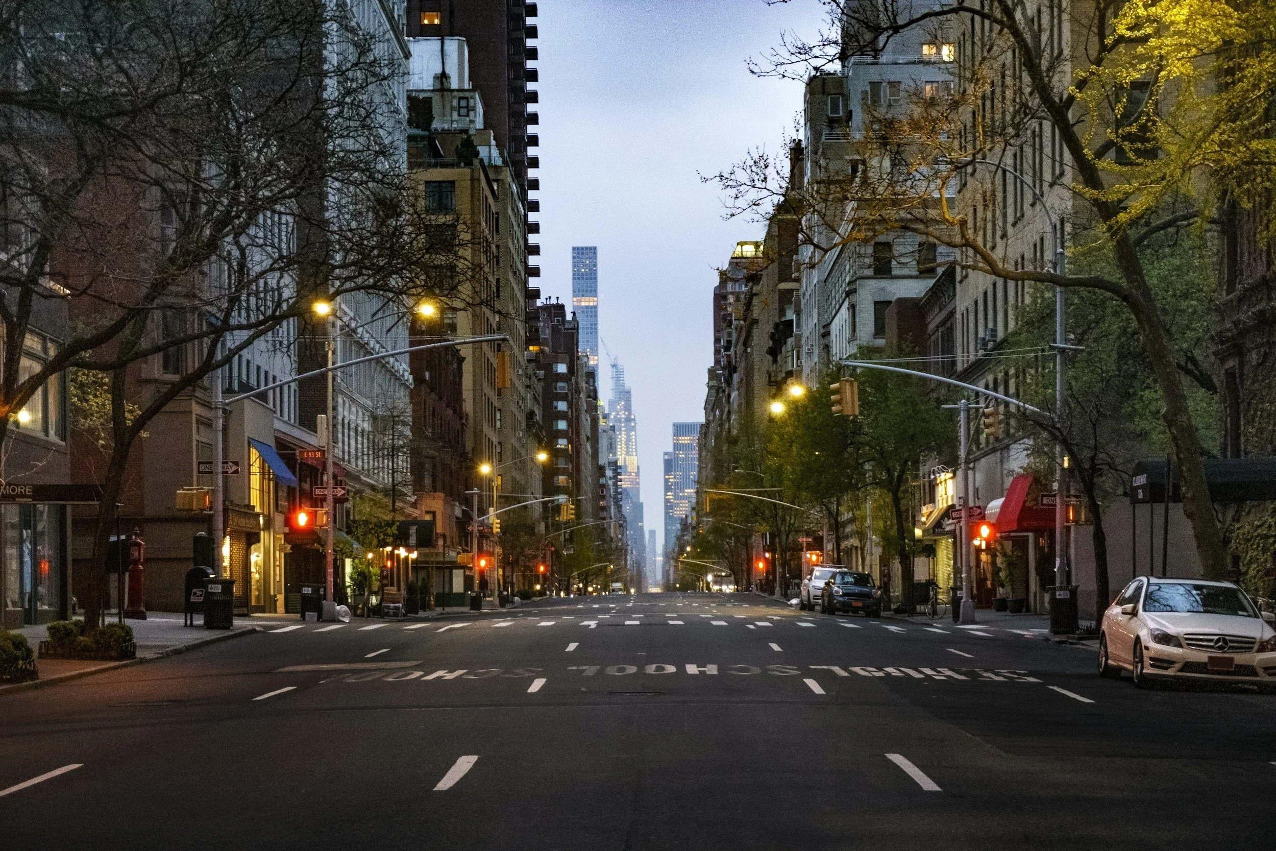 An empty city street at dusk with buildings on both sides, streetlights on, and a view of tall skyscrapers in the distance.