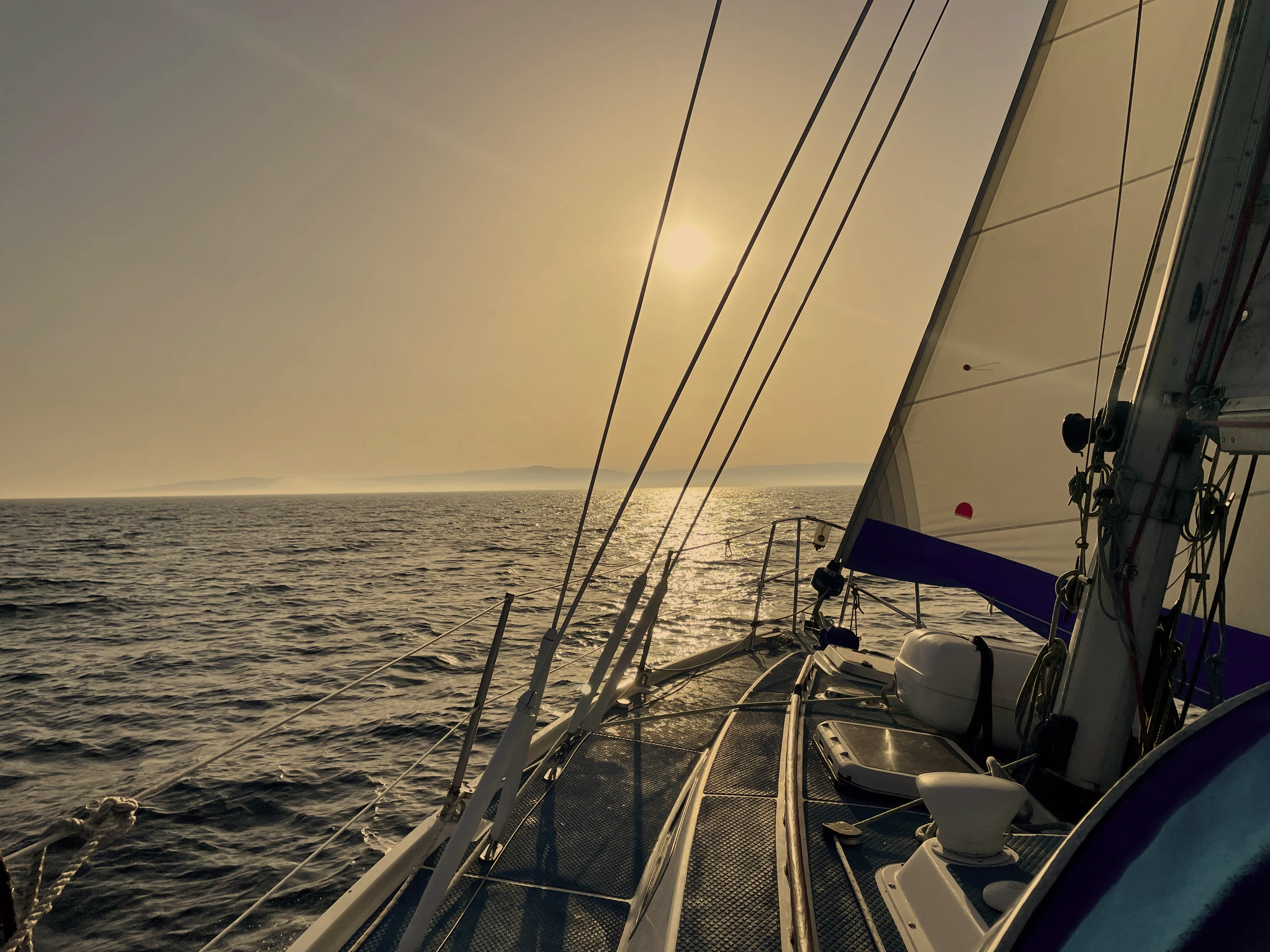View from a boat at sunset showing the ocean, with a sailboat mast and rigging in the foreground, and distant landforms across the water.