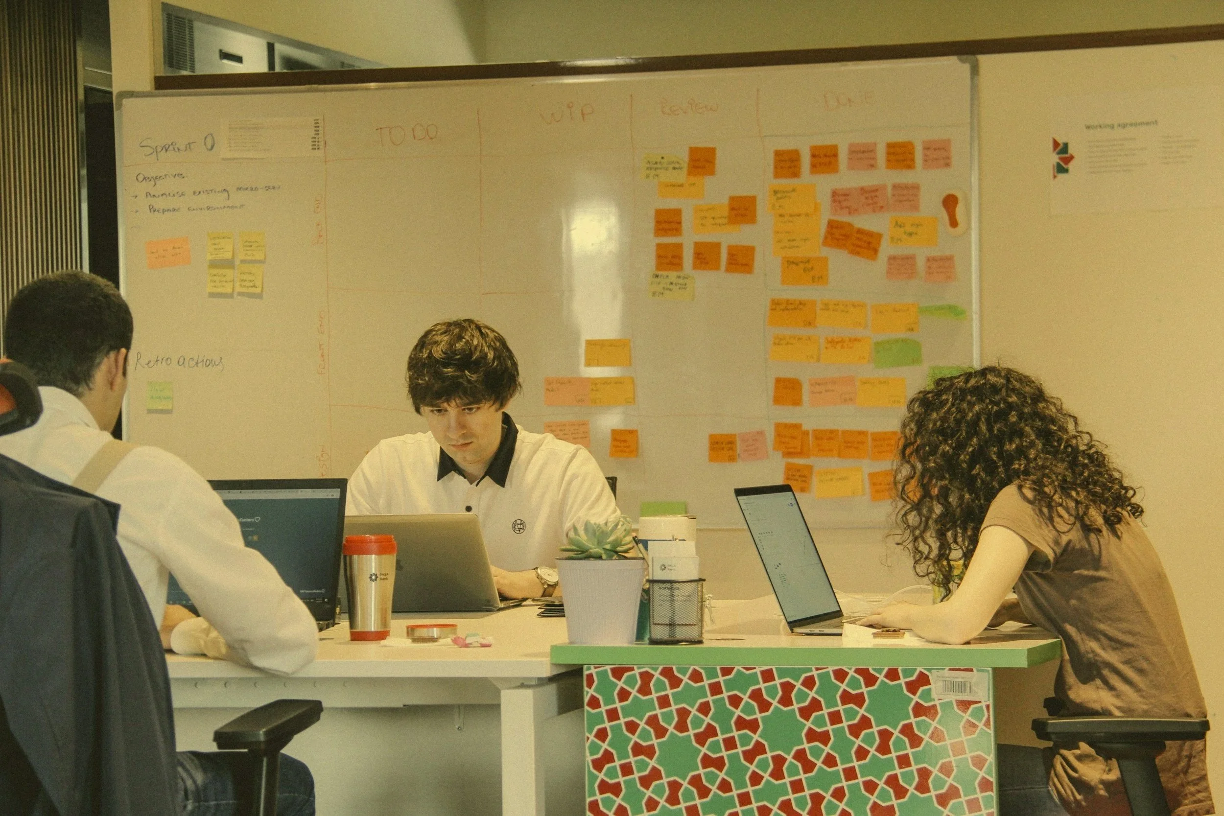 A group of people sitting at a table in a meeting or office environment, working on laptops and tablets, with a whiteboard in the background covered in sticky notes and writing.