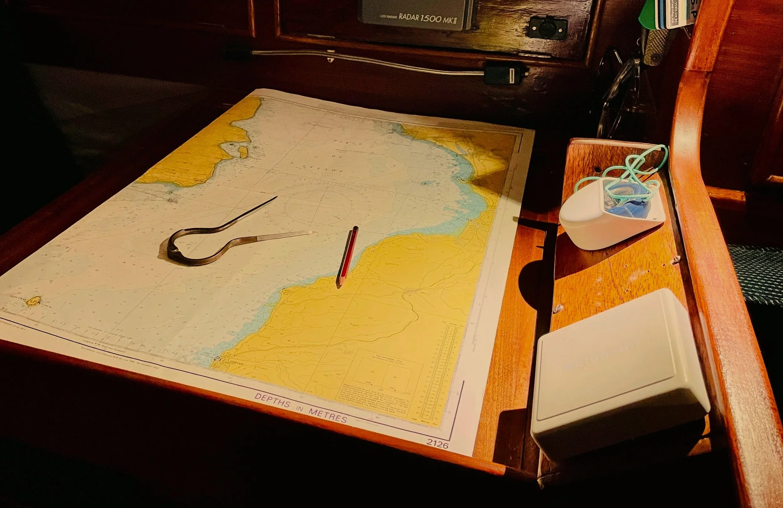 A nautical chart with a pair of tweezers, a pencil, and a marker on top, on a wooden surface, with a desk organizer holding cables and a small plastic container with rubber bands.
