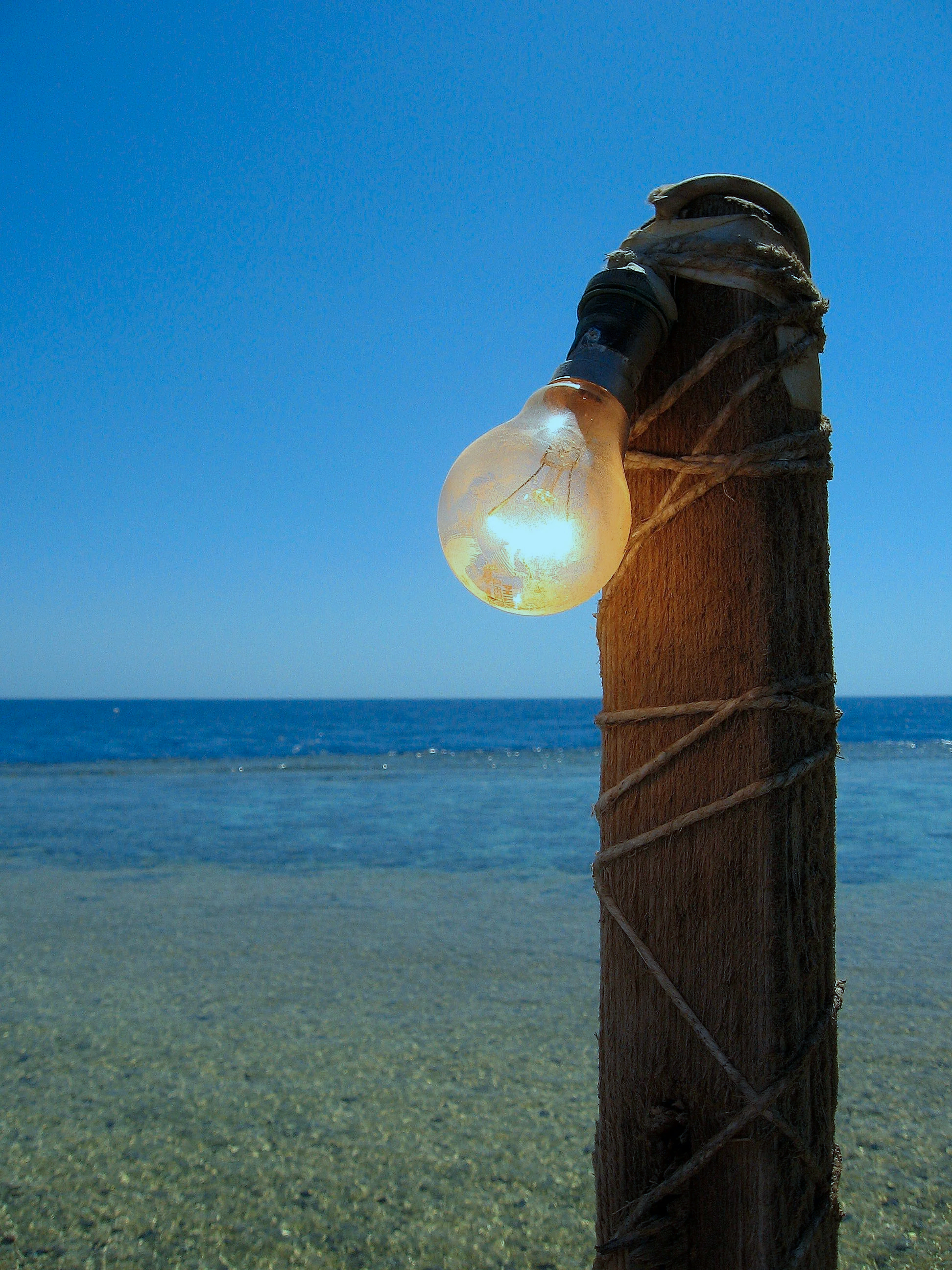 A light bulb attached to a wooden post with rope, set against a background of blue sky and ocean.