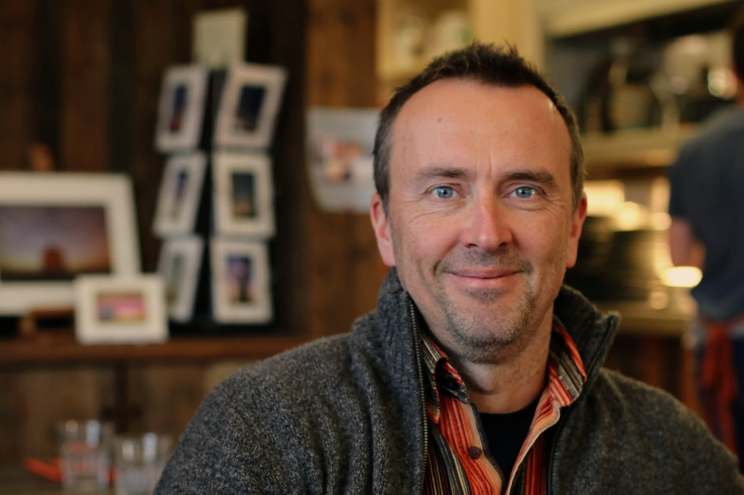 A middle-aged man with blue eyes and short dark hair smiling while sitting in a warmly lit indoor space with framed photos and shelves in the background.