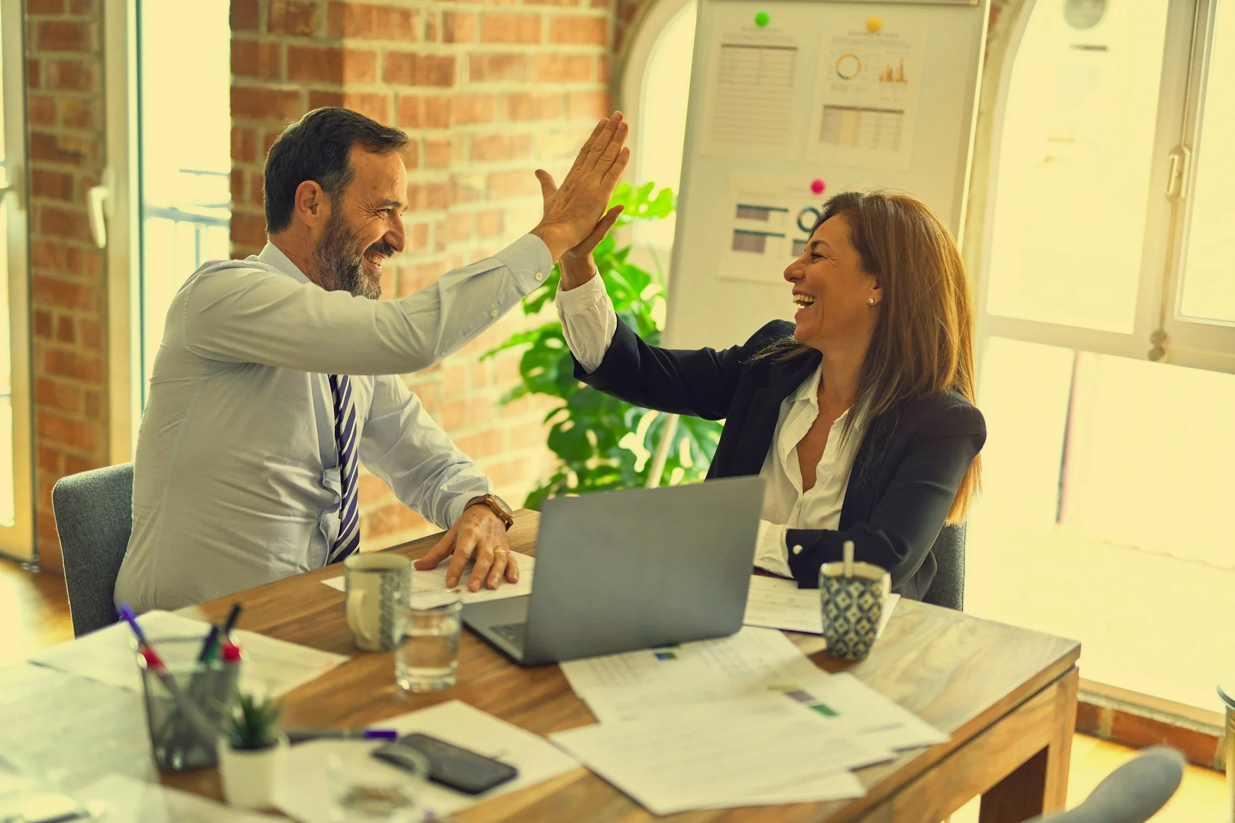 Two professionals, a man and a woman, smiling and giving each other a high five in an office setting. The man has a gray beard and is wearing a white shirt with a striped tie. The woman has long brown hair and is dressed in a black blazer and white blouse. The desk has papers, a laptop, coffee cups, and office supplies, with a brick wall and window in the background.
