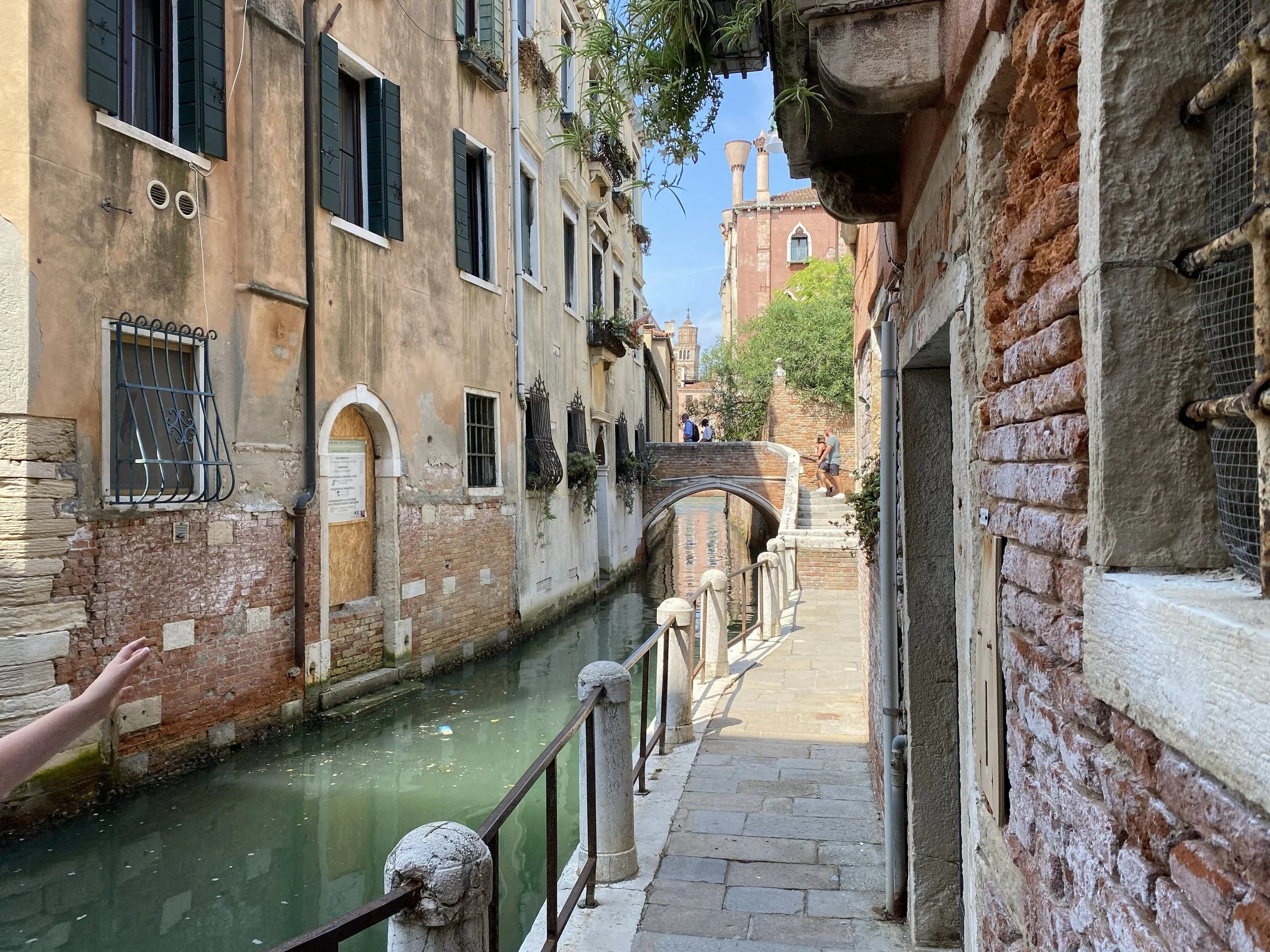 A narrow canal lined with old European buildings, some with black shutters and small balconies, a small bridge crossing the canal, with two pedestrians walking on it, and a bright blue sky visible.