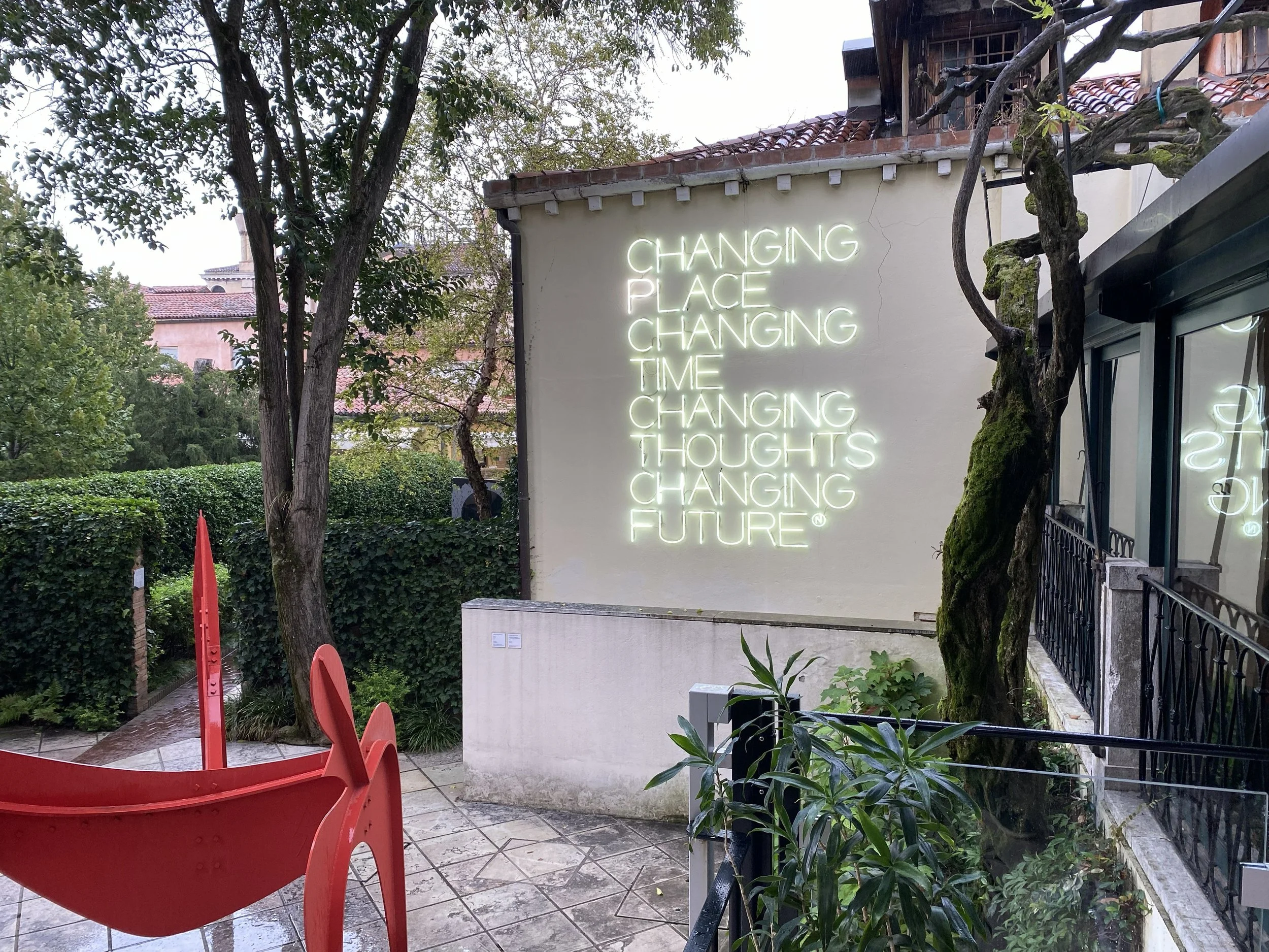 An outdoor seating area with red chairs, green plants, trees, and a white wall with a neon sign that reads "Changing place changing time changing thoughts changing future."