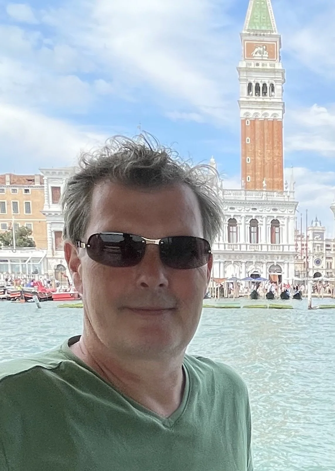 A man wearing sunglasses and a green shirt standing in front of water with the St. Mark's Campanile in Venice in the background.