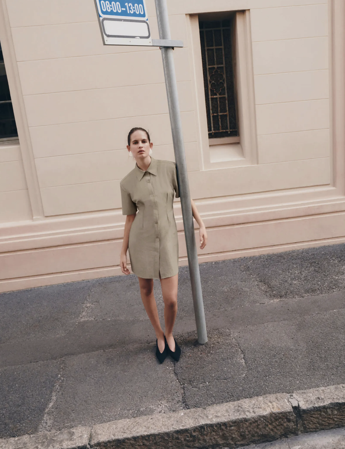 A woman with dark hair tied back, wearing a beige knee-length dress and black high heels, standing on the sidewalk next to a street sign and a beige building with a window and decorative grille.