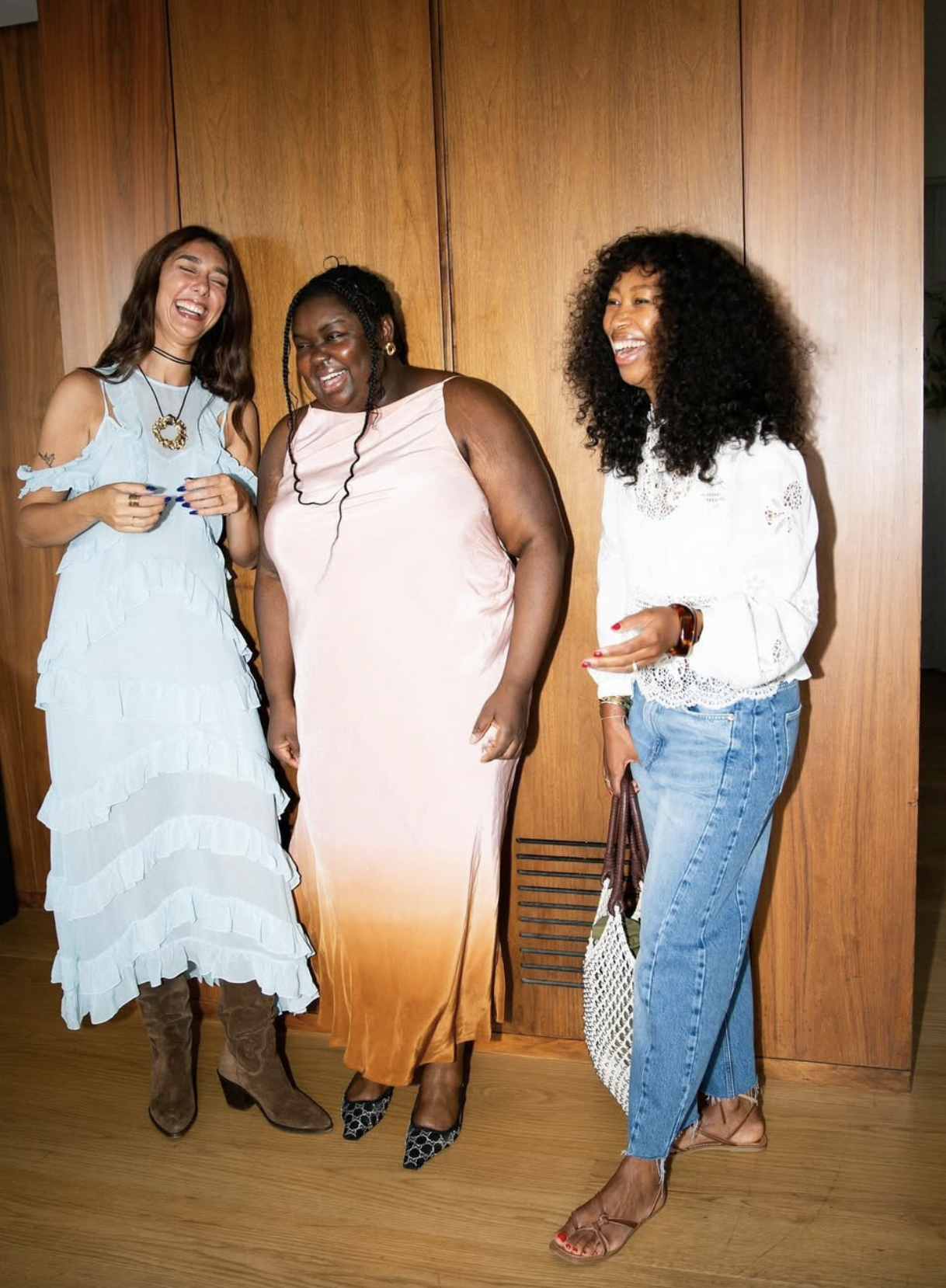 Three women standing against a wooden wall, laughing and smiling, dressed in casual and semi-formal attire.