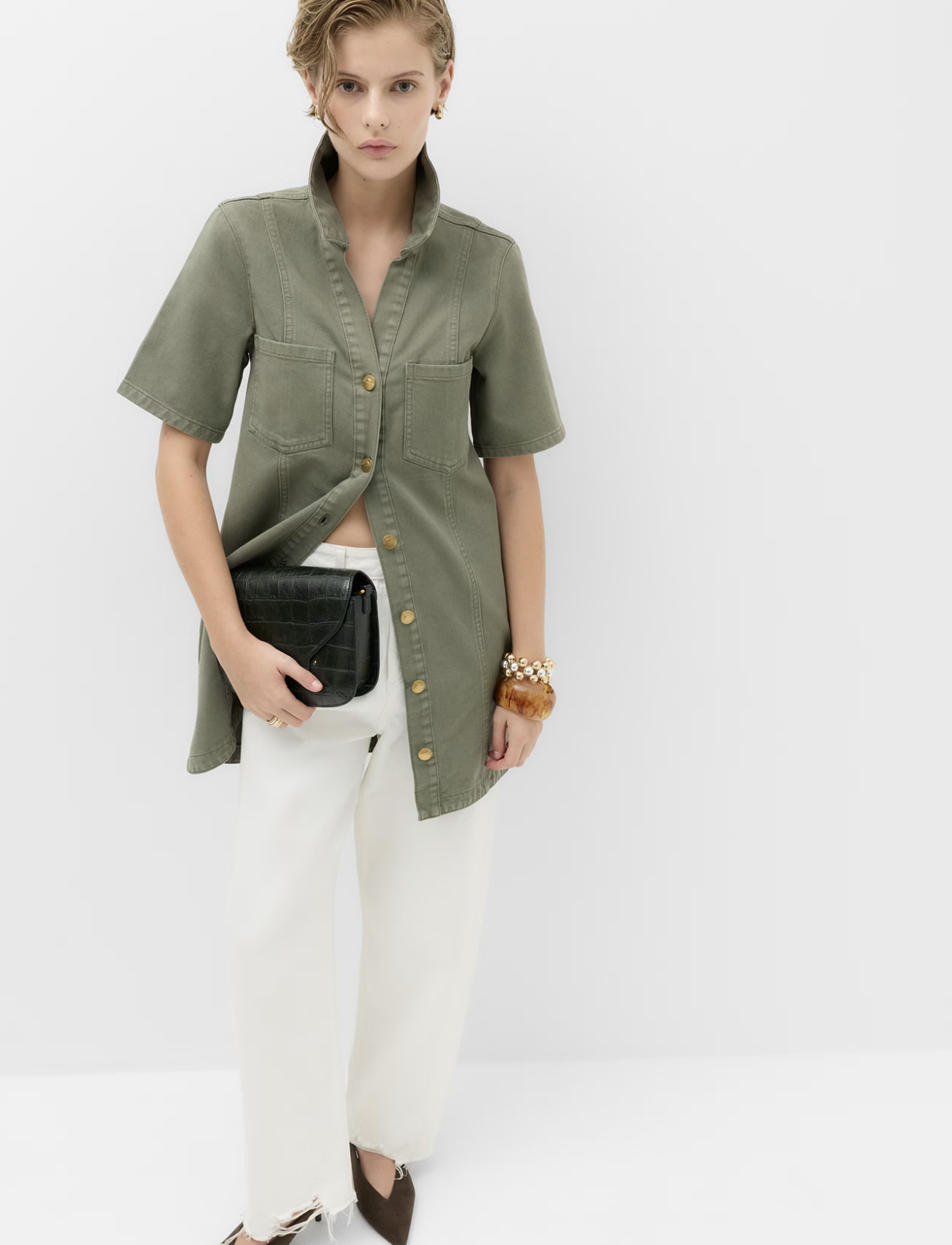 A woman with short blonde hair and earrings is wearing a green button-up shirt dress, white pants, and brown shoes, holding a black clutch bag, with a bracelet on her left wrist, against a plain light background.