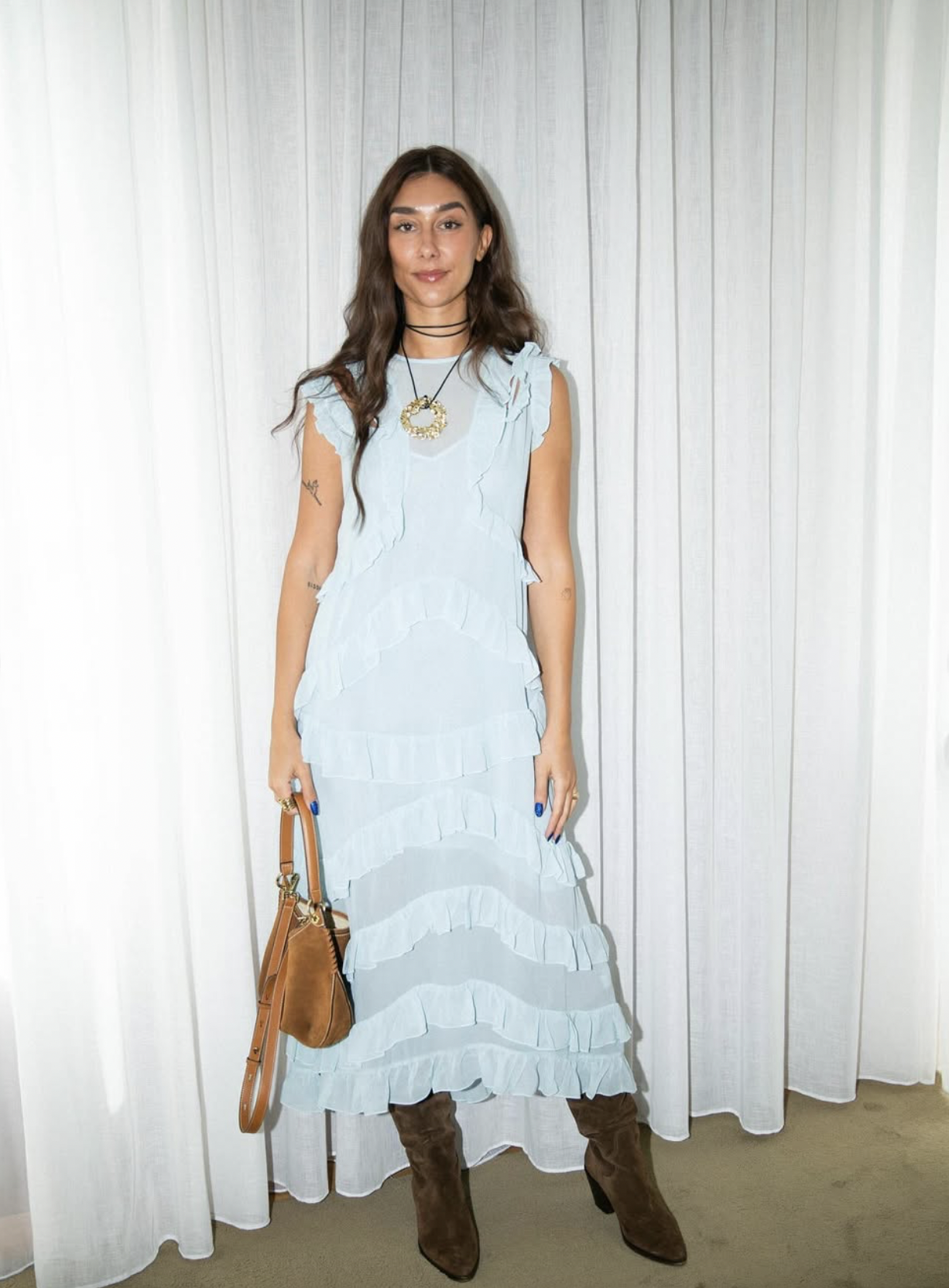 A young woman with long brown hair wearing a light blue ruffled maxi dress, brown boots, and holding a tan handbag, standing in front of a white curtain.