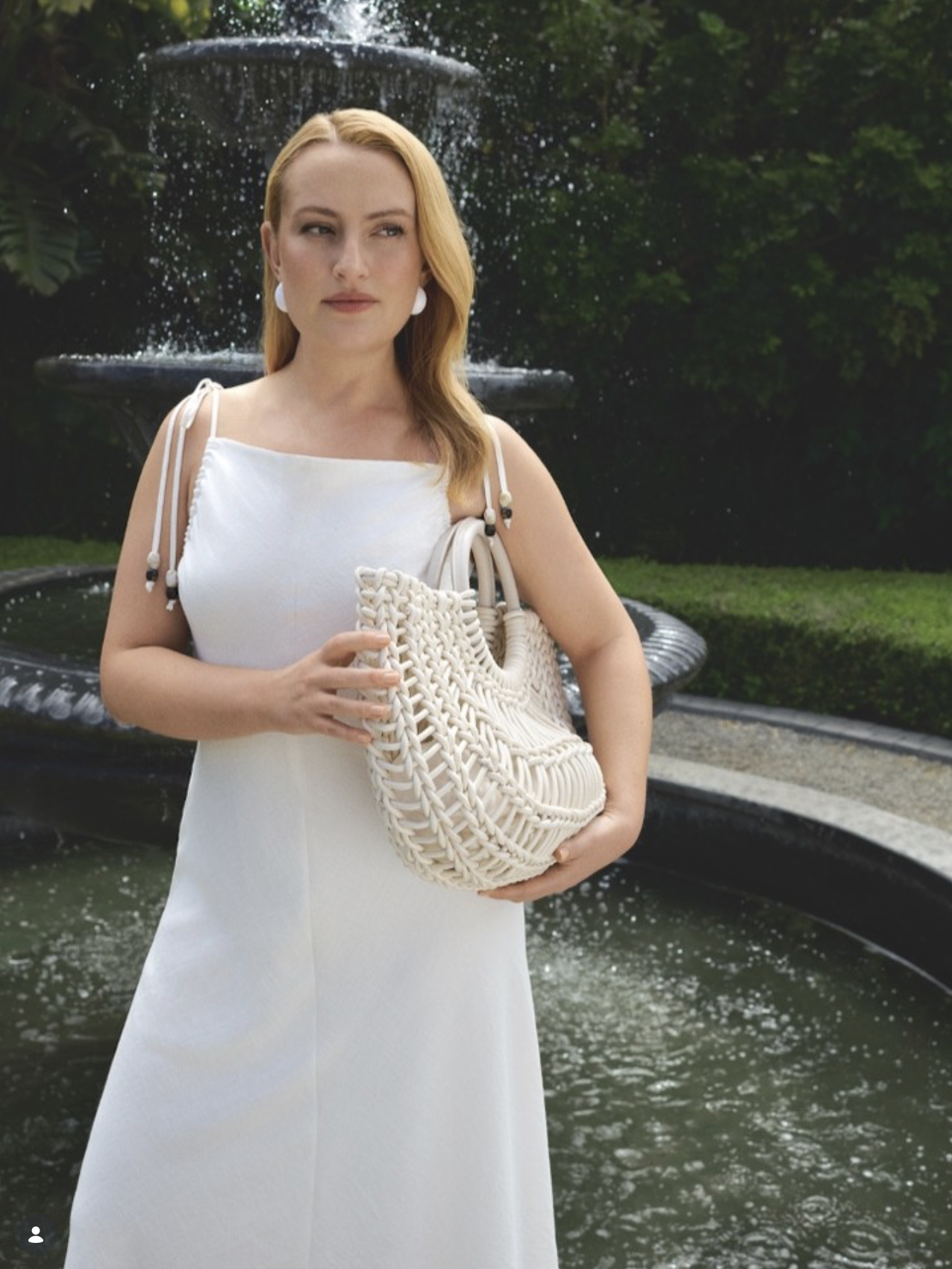 A woman with red hair wearing a white dress and earrings, holding a woven handbag stands in front of a fountain with water cascading from multiple levels, surrounded by greenery.