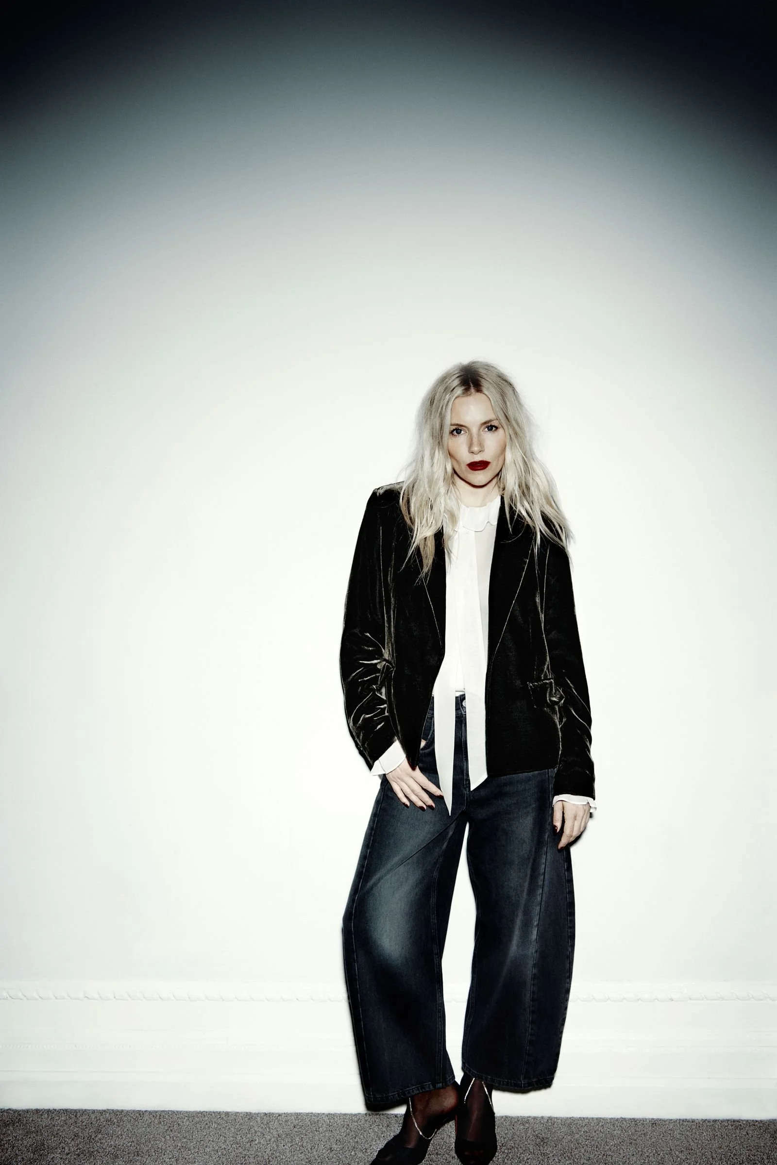 A woman with blonde, wavy hair and dark red lipstick stands with one hand on her hip against a plain white background. She is wearing a black velvet blazer, a white blouse, dark wide-leg jeans, and black heels.