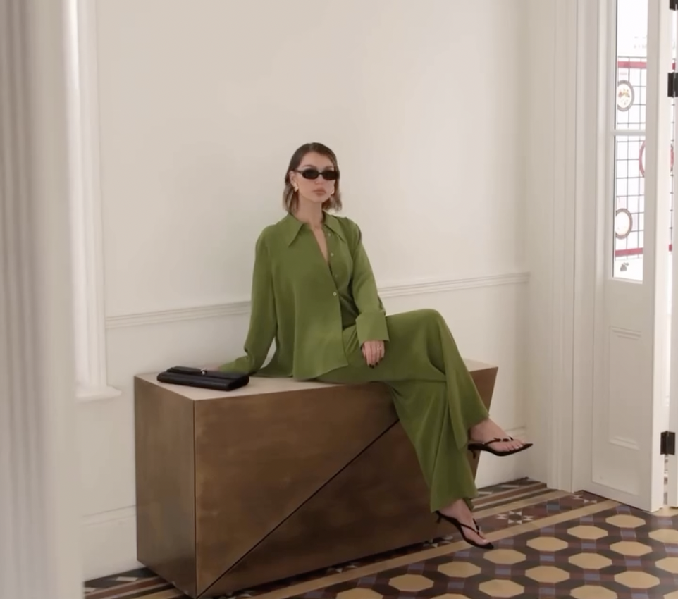 Woman in green outfit and sunglasses sitting on a wooden bench in a well-lit room with patterned tile floor and glass-paneled door.