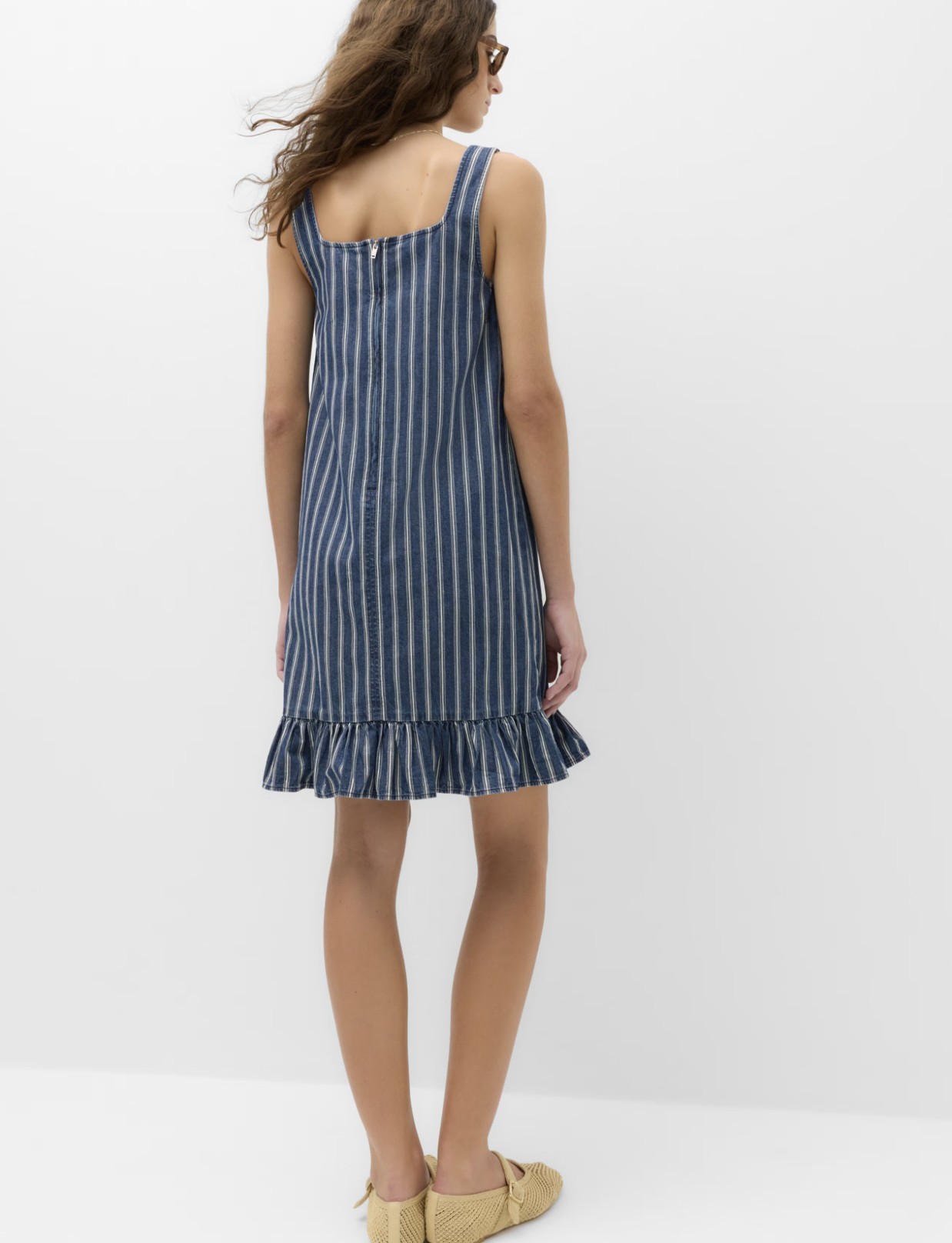 A woman with wavy brown hair wearing sunglasses and a sleeveless blue striped dress with a ruffled hem, standing against a plain white background, showing the back of the dress.