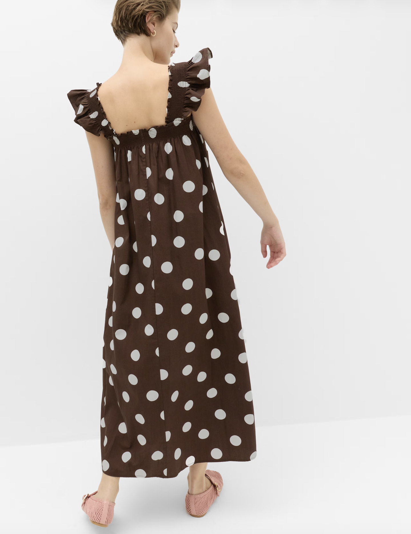 A woman wearing a brown polka dot dress with ruffled sleeves and pink shoes standing against a plain white background.