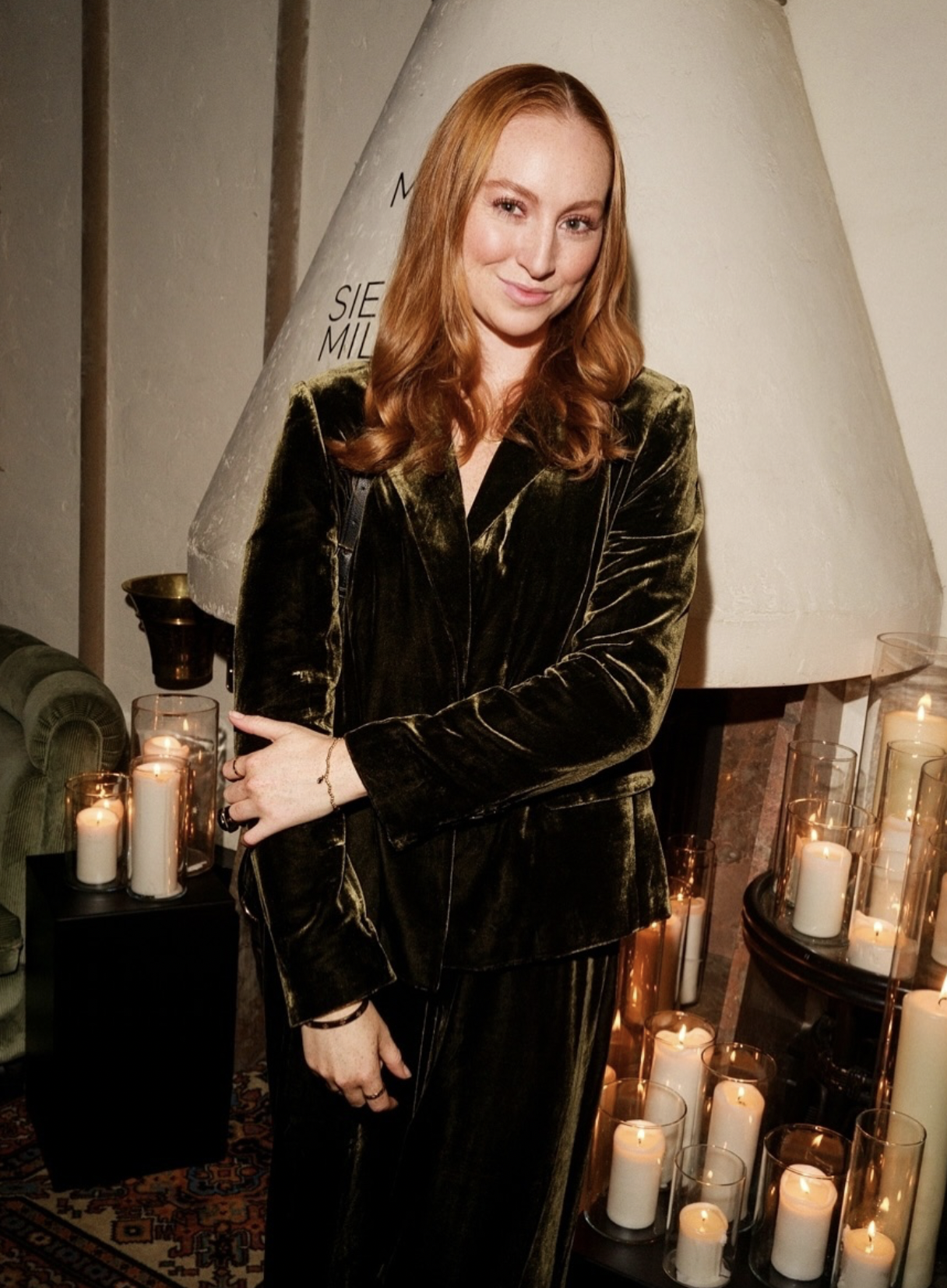 A young woman with red hair wearing a black velvet suit standing indoors near a fireplace with lit candles, posing with her arms crossed and smiling slightly.
