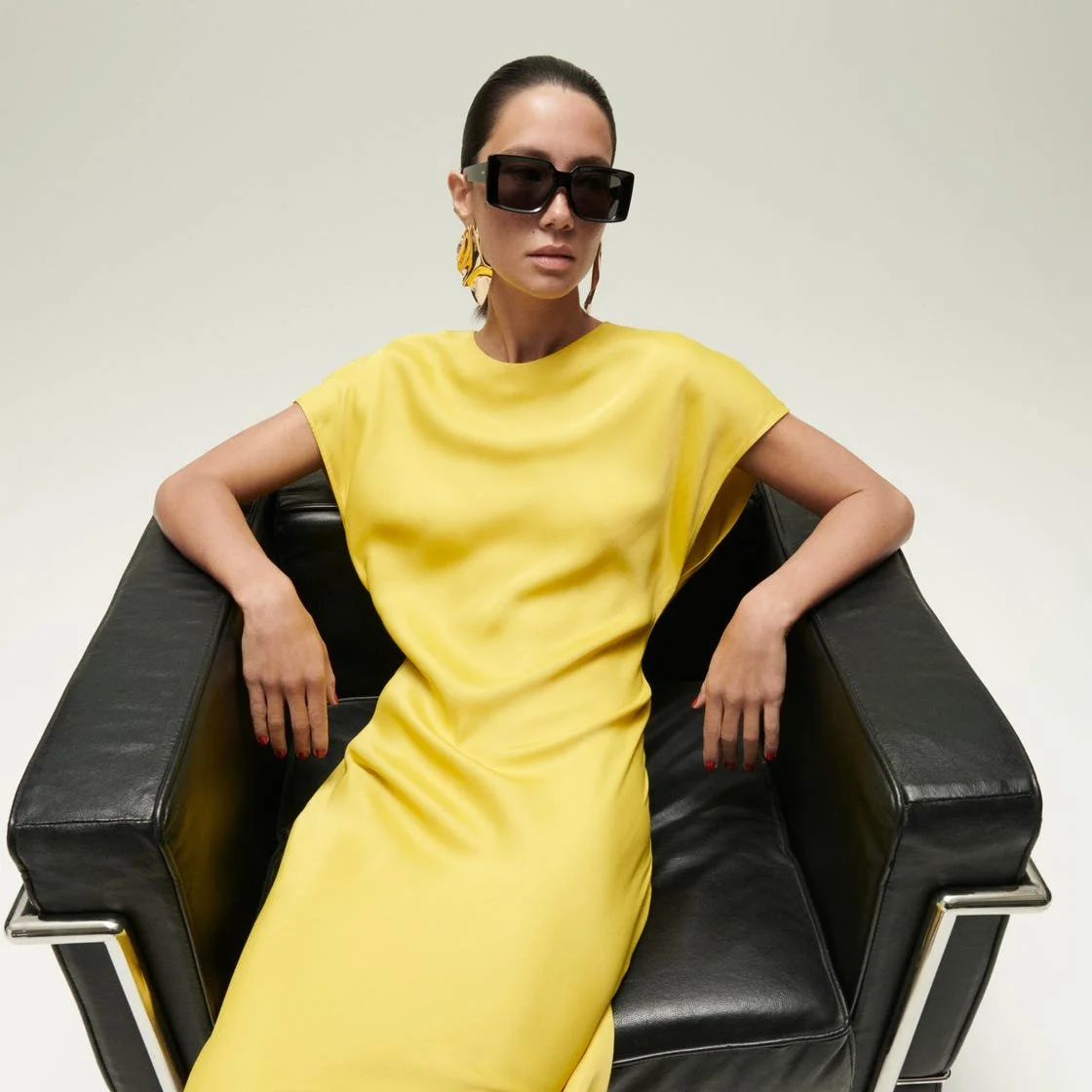A woman wearing a yellow dress, large black sunglasses, and gold earrings sitting on a black leather chair against a plain background.
