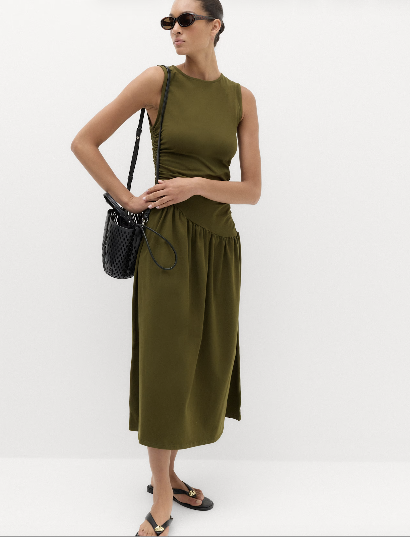 A woman wearing an olive green sleeveless dress, black sandals with gold accents, and sunglasses, standing against a plain white background.