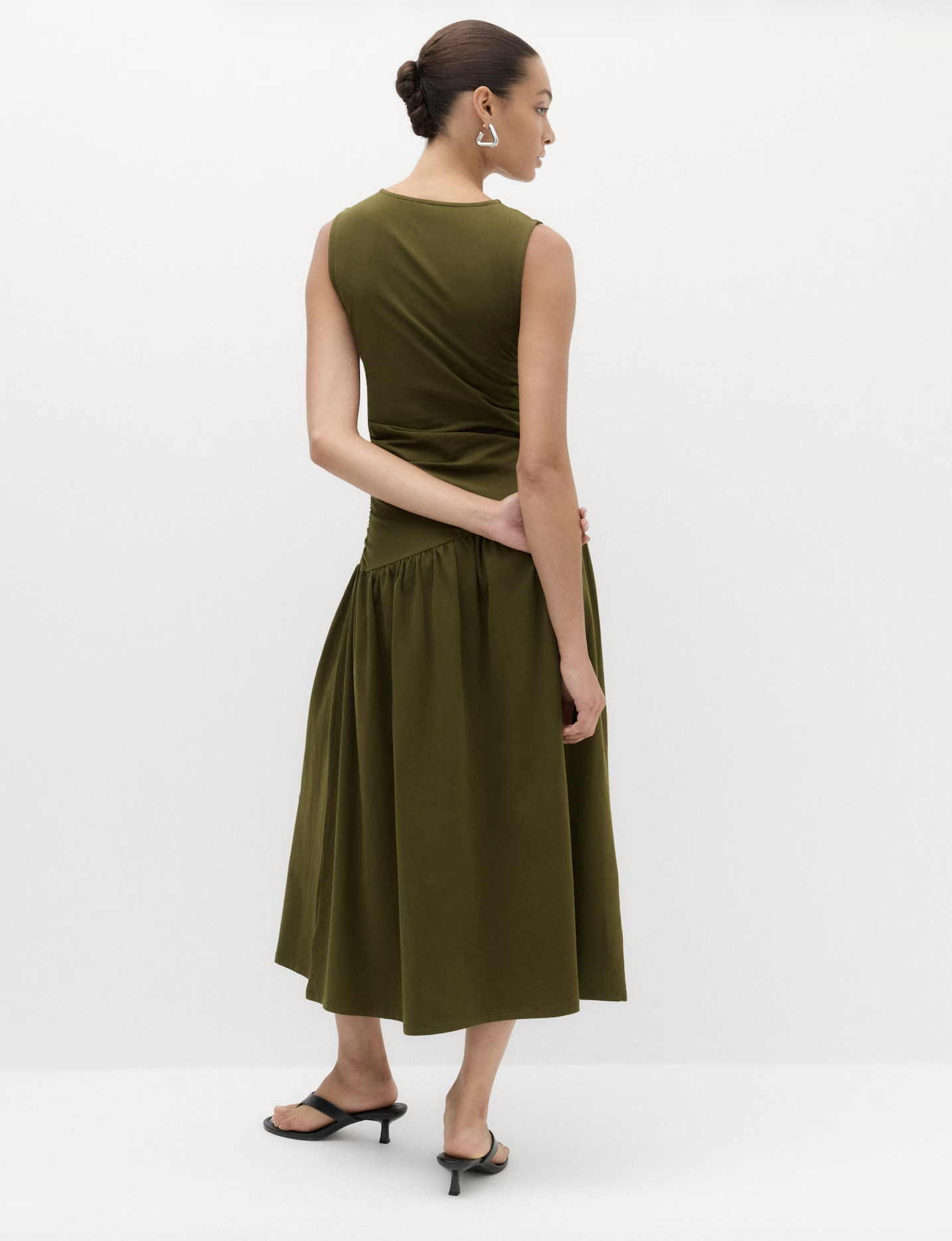 Woman with brown hair styled in a bun, wearing an olive green sleeveless dress with a full-length skirt, black high-heeled sandals, and silver earrings, standing against a plain white background.