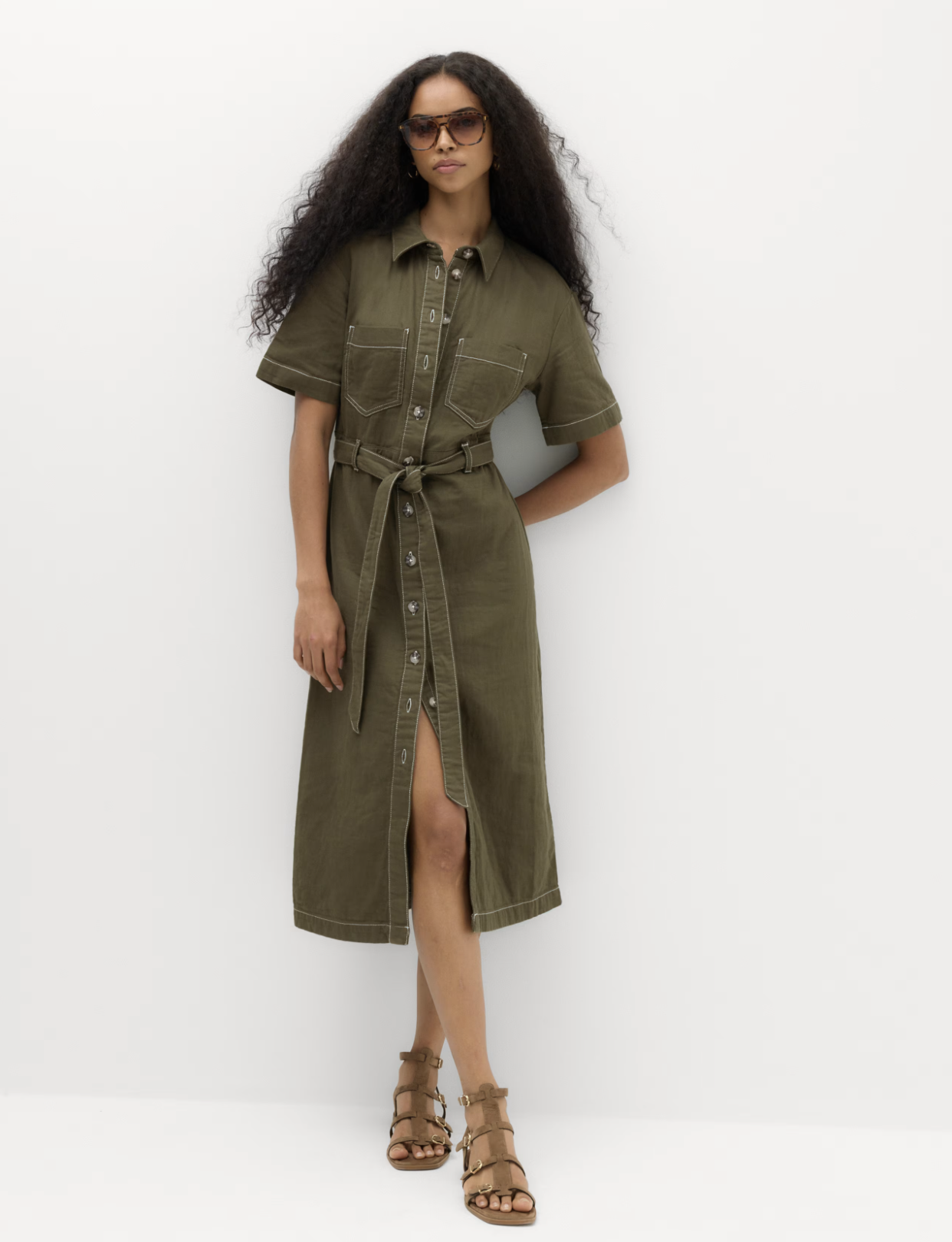 Woman wearing an olive green button-up shirt dress with short sleeves, in front of a plain white background.