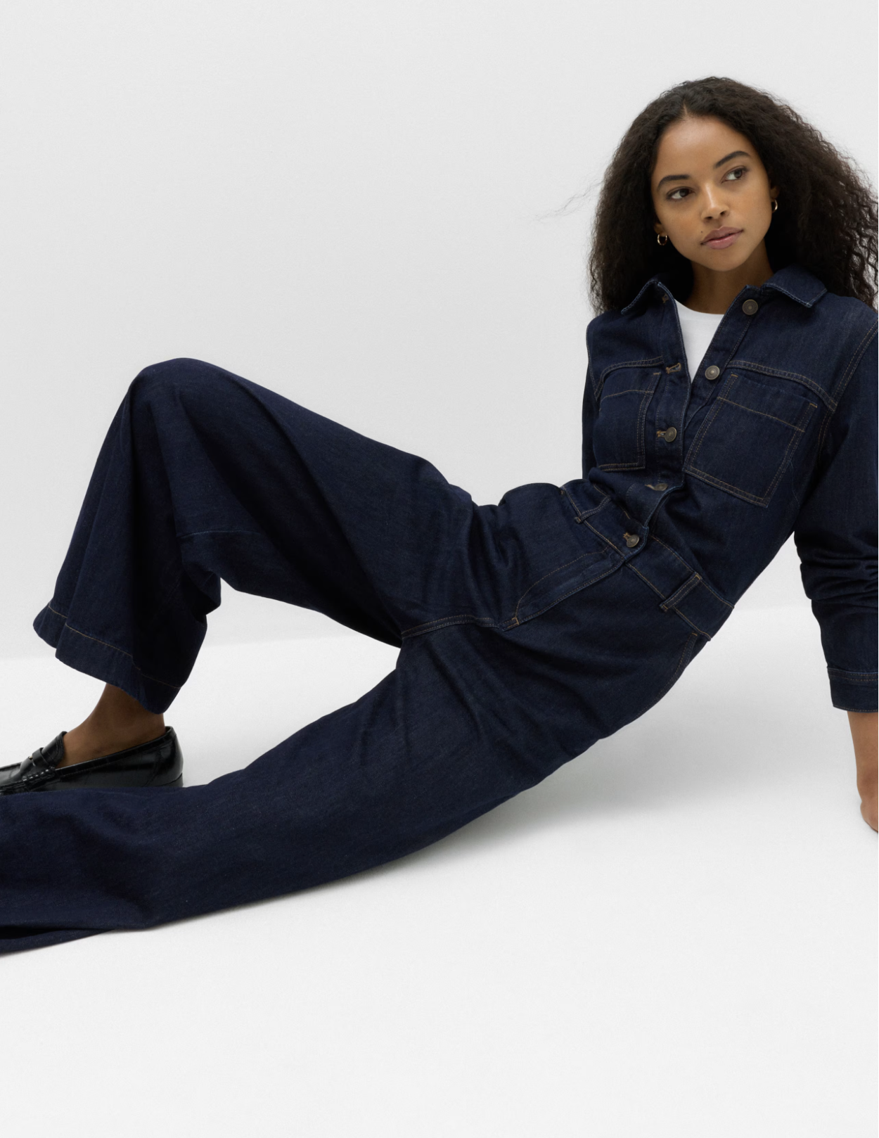 Young woman with curly hair wearing a dark denim jacket and pants, black loafers, sitting on a white background.