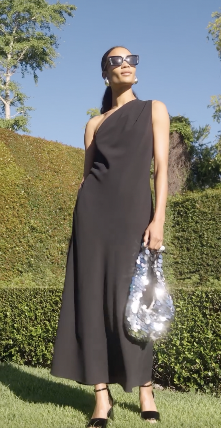 A woman in a sleeveless black dress with sunglasses, standing outdoors in a garden, holding a silver sequin bag, wearing high heels, and looking off into the distance.