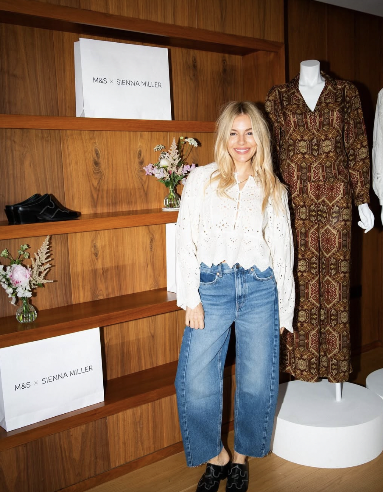 A woman with blonde hair smiling at a fashion event or boutique, standing in front of wooden shelves and a mannequin dressed in a patterned dress. There are flowers on the shelves and a sign that reads "M&S x Sienna Miller".