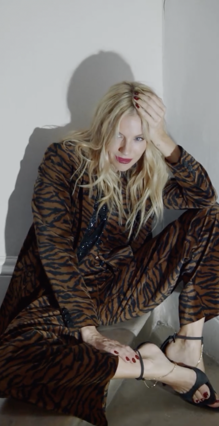 Blonde woman in a tiger print outfit sitting on the floor against a white wall, holding her head with one hand, with a serious expression.