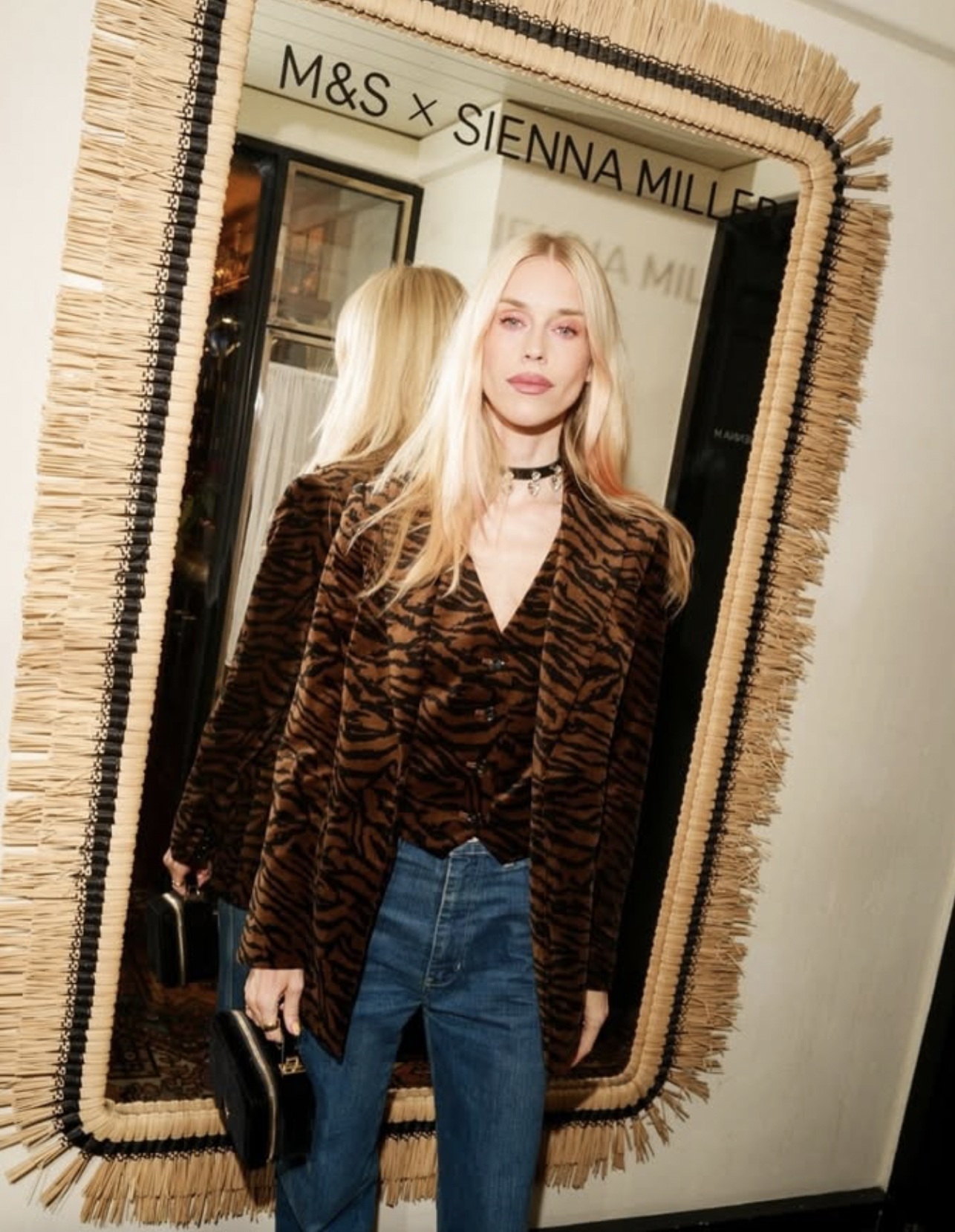 A woman with blonde hair wearing a tiger-striped blazer and blue jeans is taking a selfie in a mirror. The mirror has a woven frame, and there are signs for "M&S x SIENNA MILLER" reflected in the background.