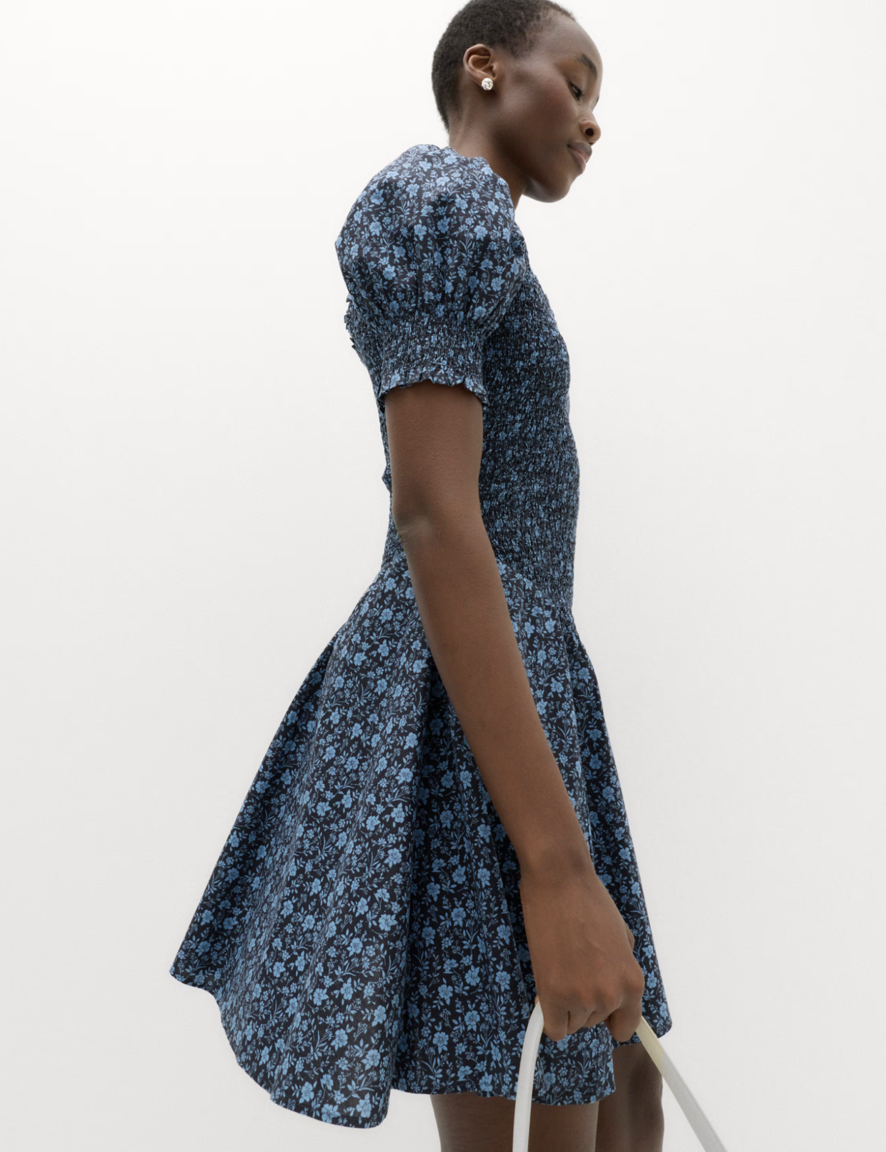 A young woman in a blue floral dress holding a white bag, standing against a plain white background. She has short hair and is wearing earrings.
