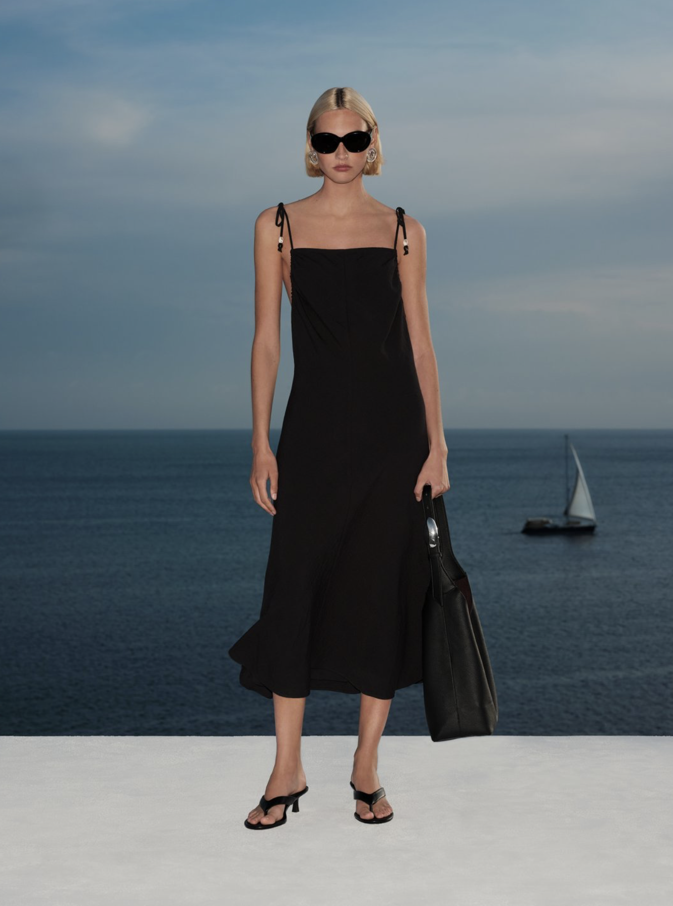 A woman in a black dress, sunglasses, and high heels stands on a white surface in front of a blue ocean with a sailboat in the distance, under a partly cloudy sky.