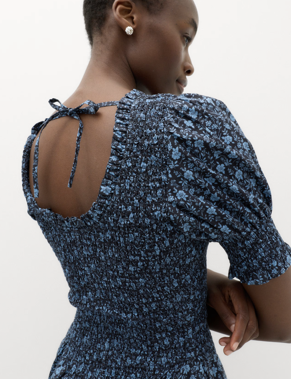 A woman with short hair and dark skin is wearing a blue floral dress with puffed sleeves and a tied back detail, standing against a plain white background.