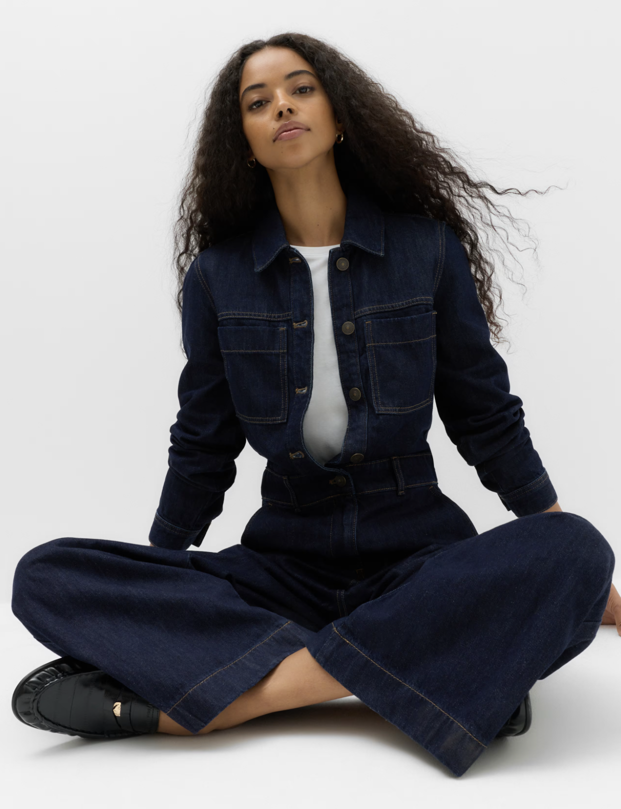 Woman sitting cross-legged on the floor, wearing a dark denim jacket and pants, with a white shirt underneath, against a plain white background.