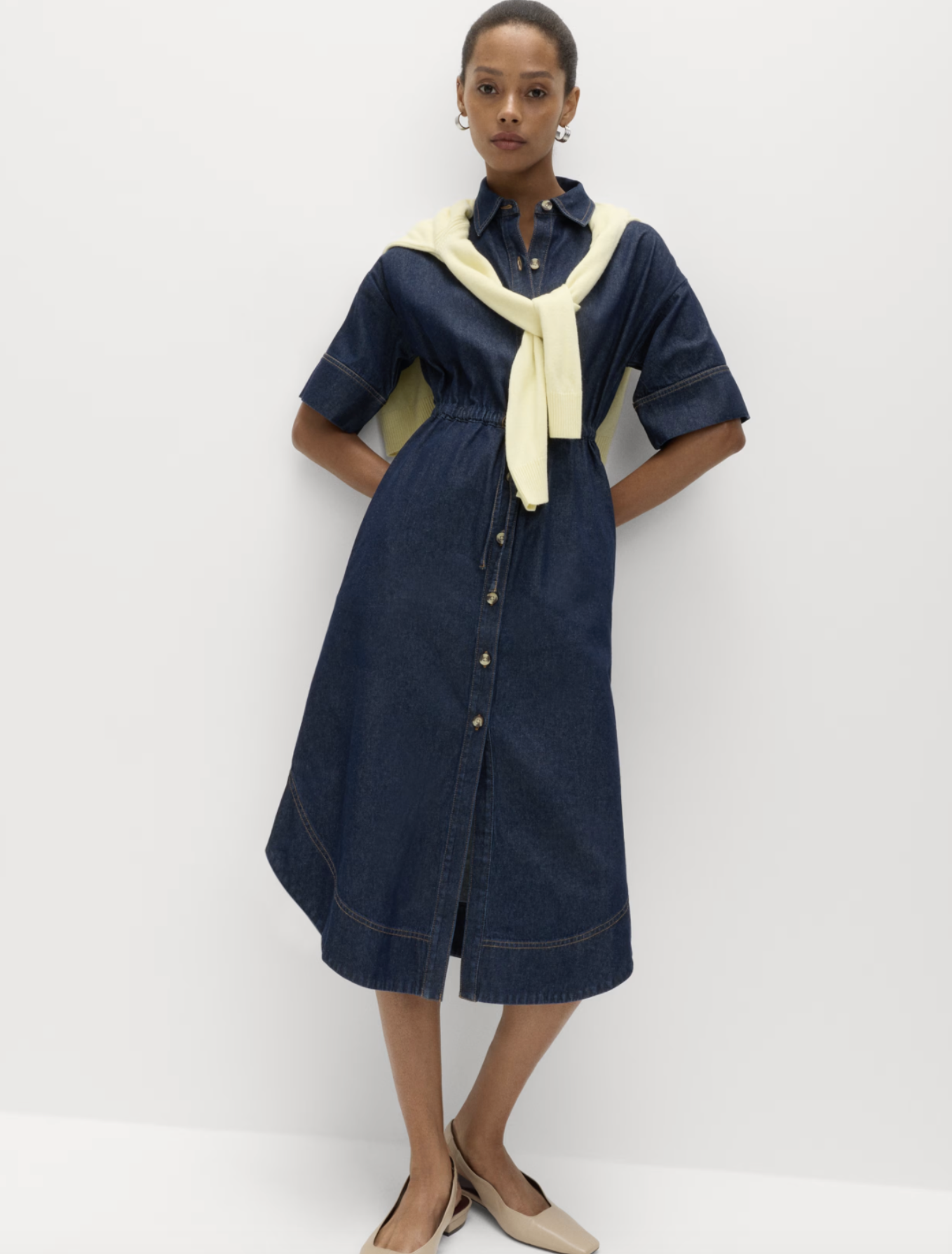A woman standing against a white background wearing a dark denim button-down dress with yellow accents, a cream-colored sweater tied around her shoulders, and beige heels.