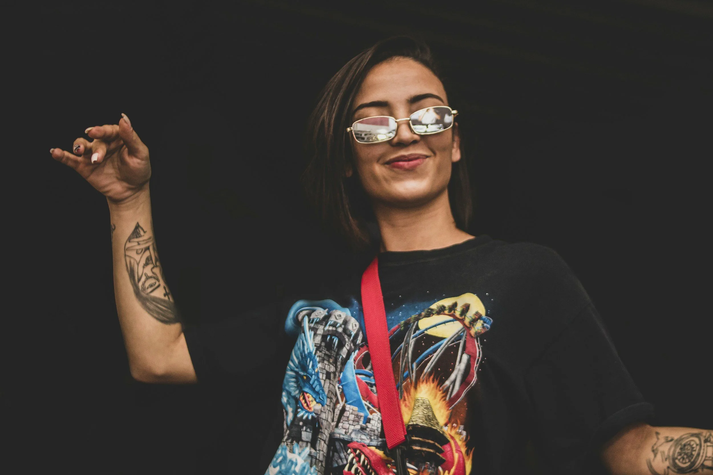 A young woman with dark hair and tattoos on her arm is smiling with her eyes closed, wearing sunglasses and a colorful graphic T-shirt, against a black background.