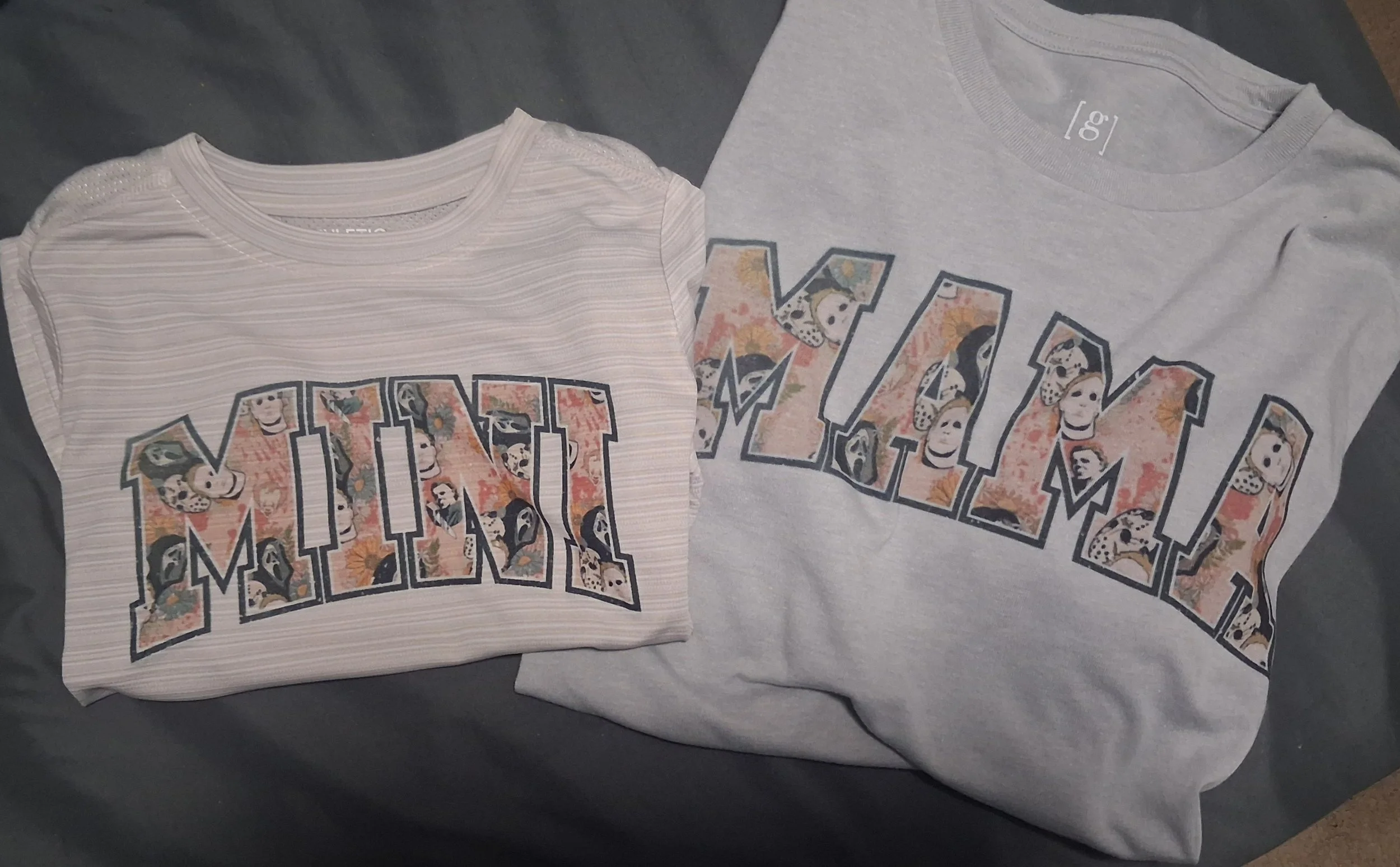 Two folded T-shirts with a printed design on top of a dark surface. The T-shirts have the same design with the word 'MOM' in bold outlined letters filled with floral and skull patterns, one shirt is light-colored with a subtle striped pattern, and the other is plain gray.