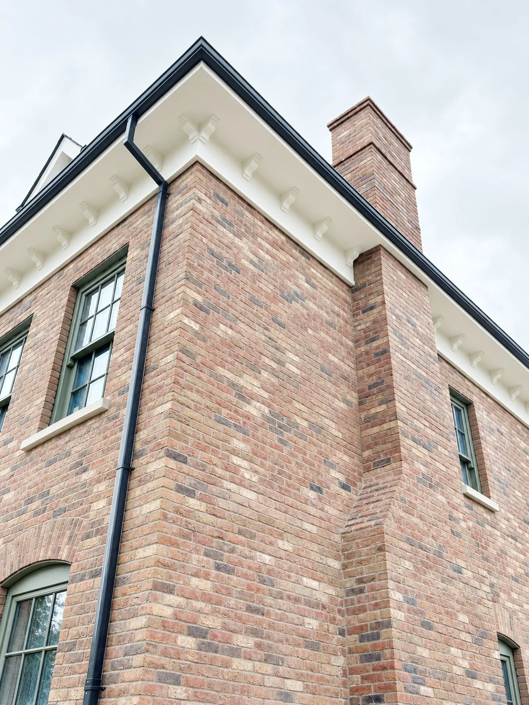 Close-up photo of a brick building corner with windows, black gutter, decorative eaves, and chimney against cloudy sky.