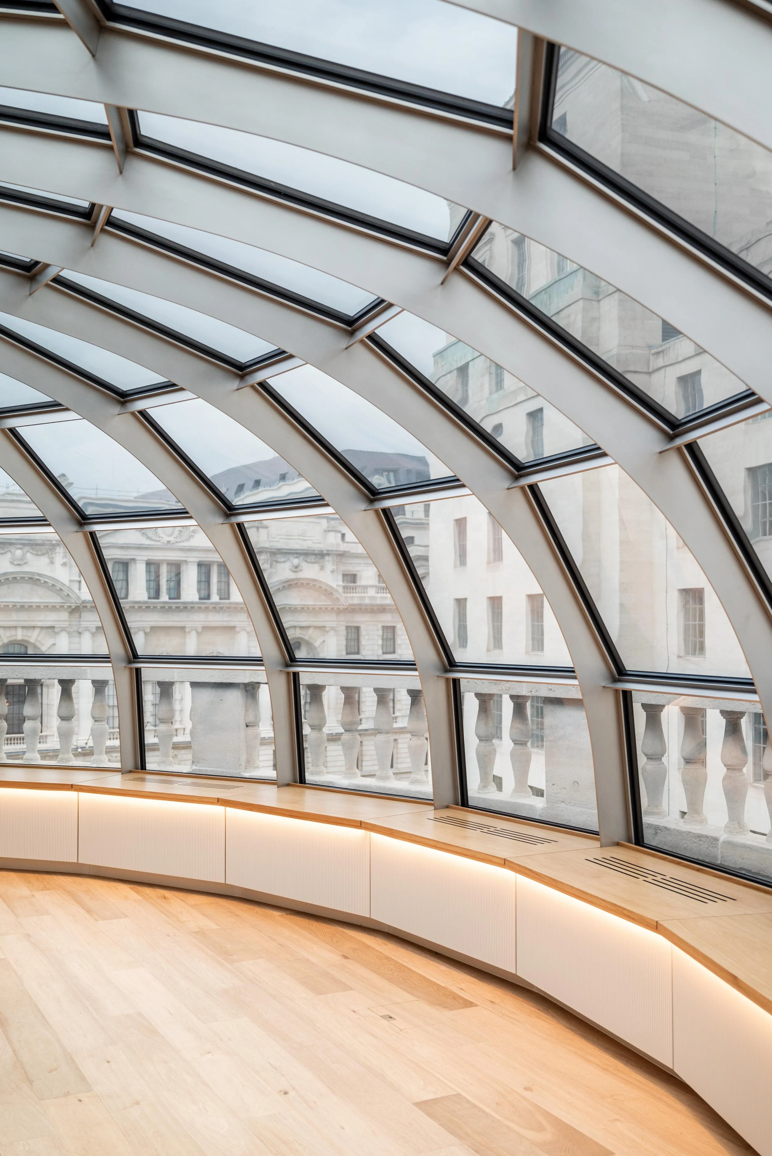 Interior view of a modern building with a curved glass ceiling, large windows, and a wooden floor. The outside view includes historic buildings with ornate facades.