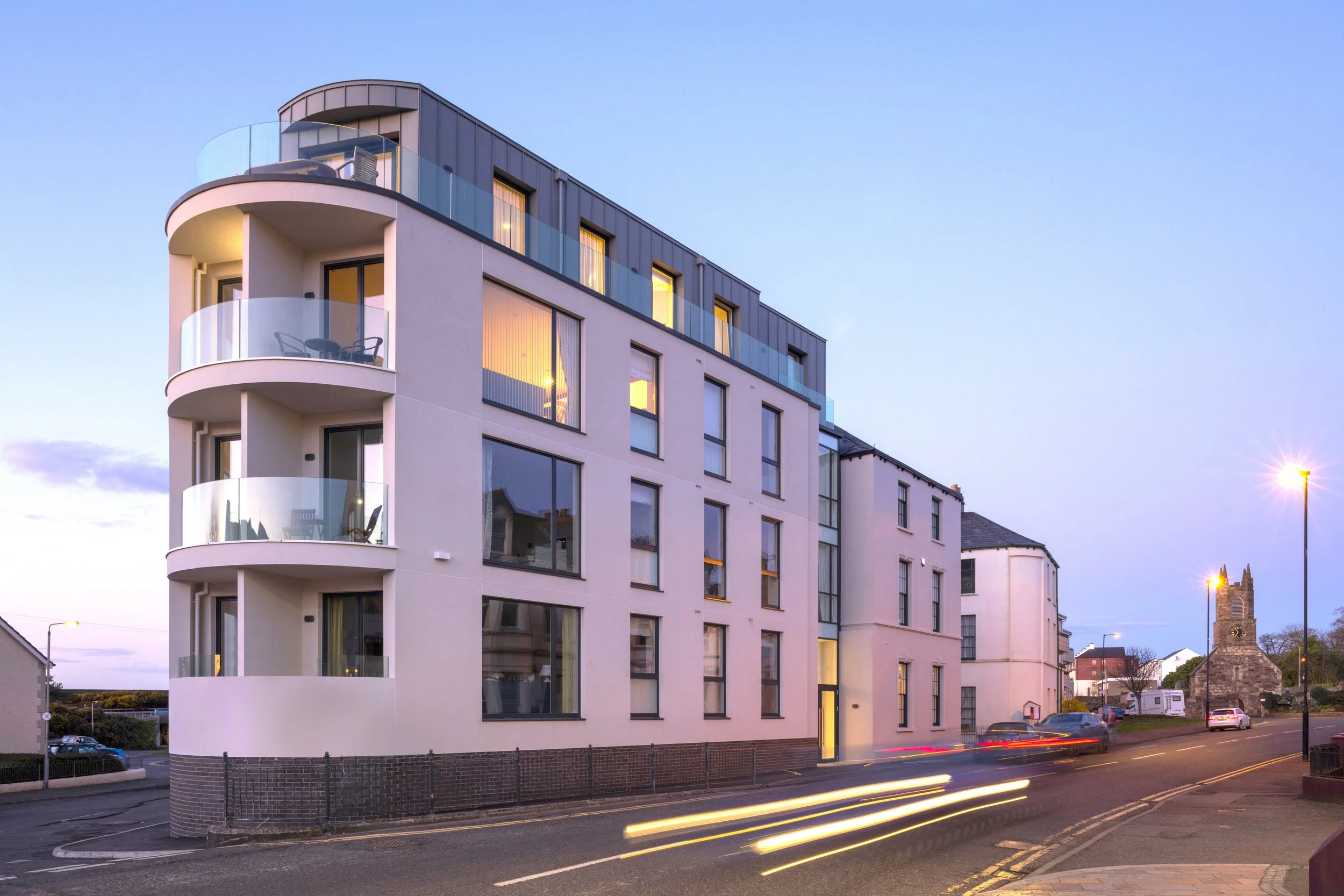 Modern multi-story residential building with curved balconies, large glass windows, and exterior lighting during dusk, with a street and passing cars in the foreground.