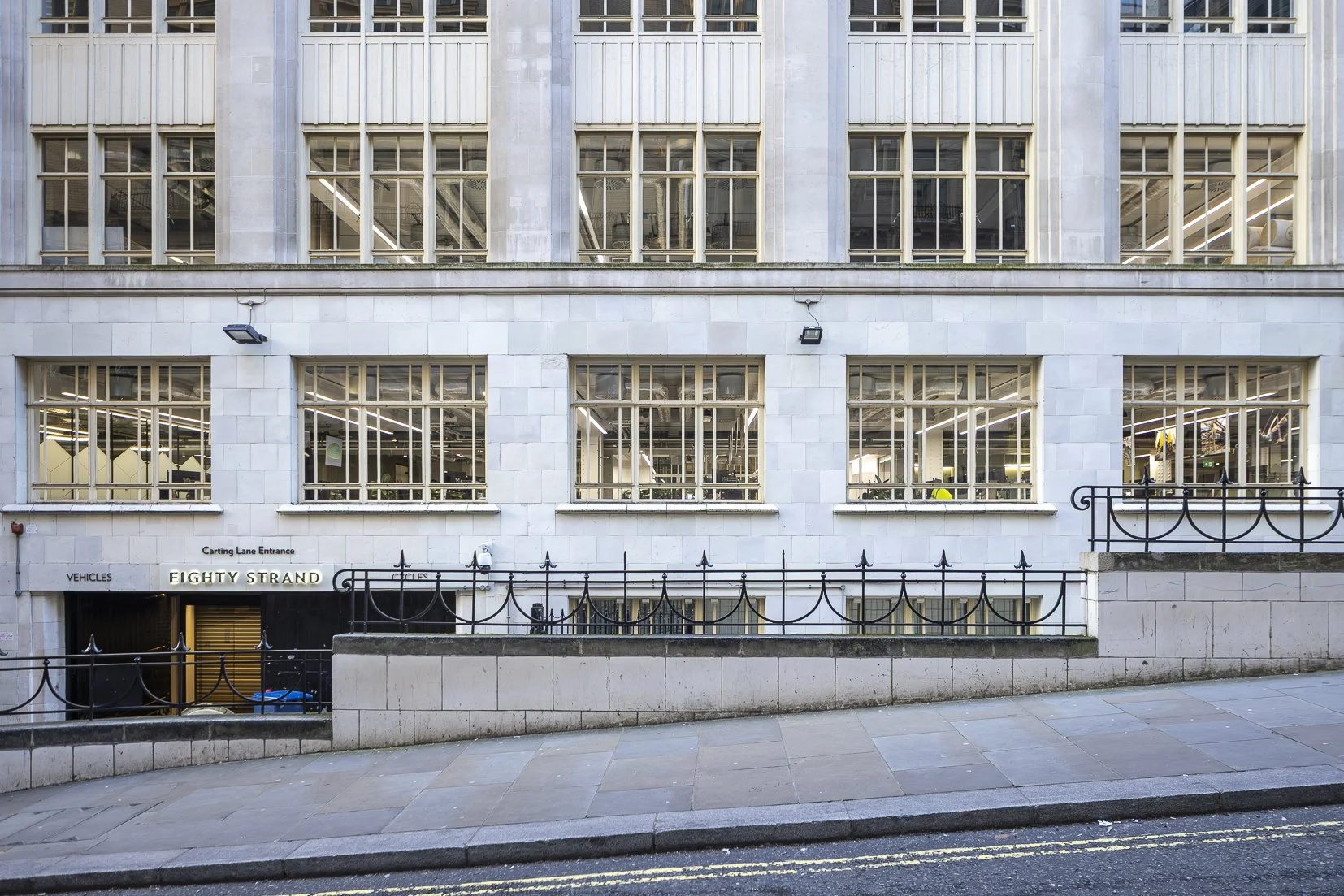 Front view of a white stone building with multiple large windows, a black wrought iron fence, and signage reading 'Eighty Strand' and 'Carting Lane Entrance'.