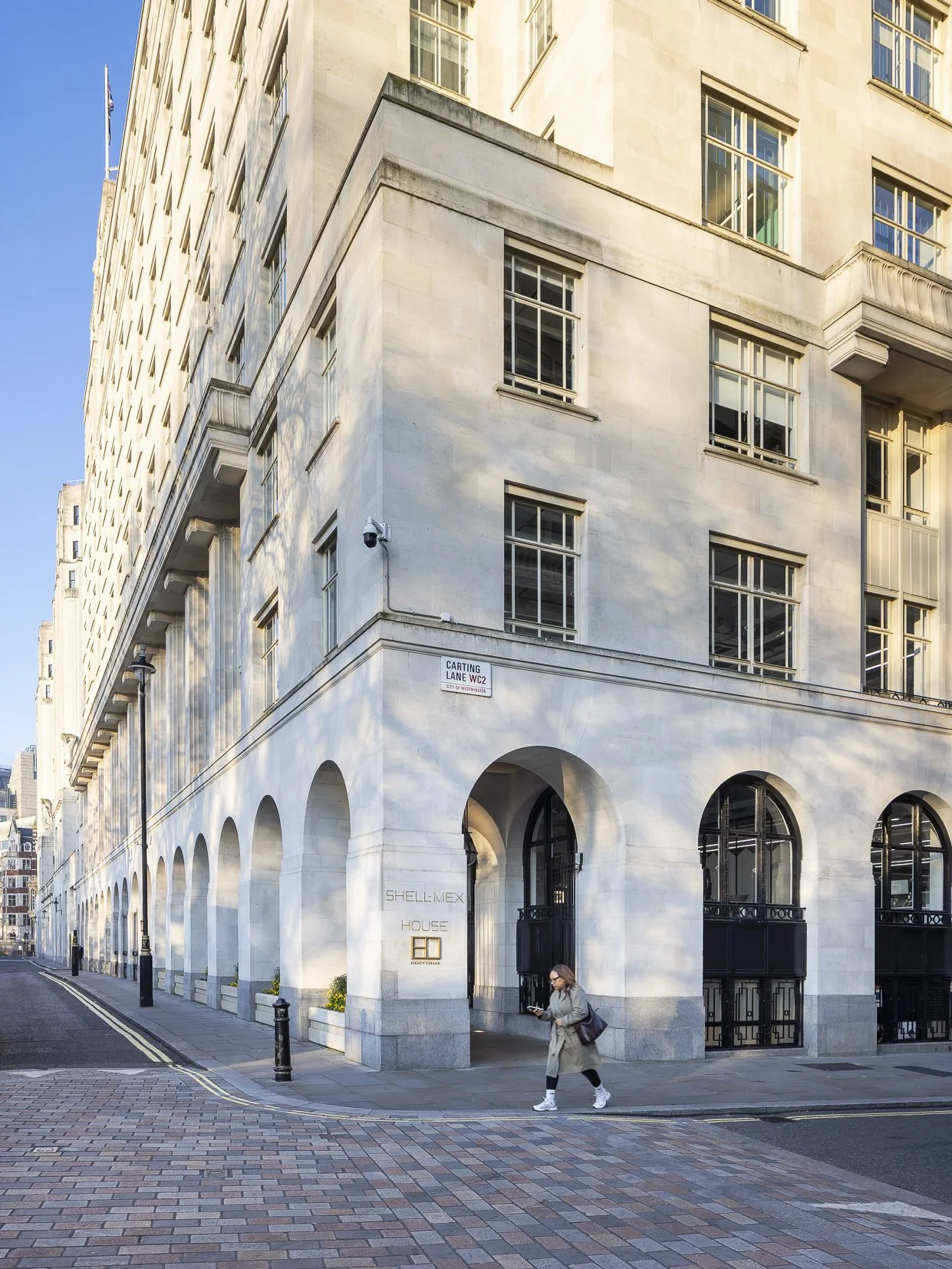 A woman walking on a street near Shellmex House, a white building with arched entrances and multiple windows, in Westminster, London.