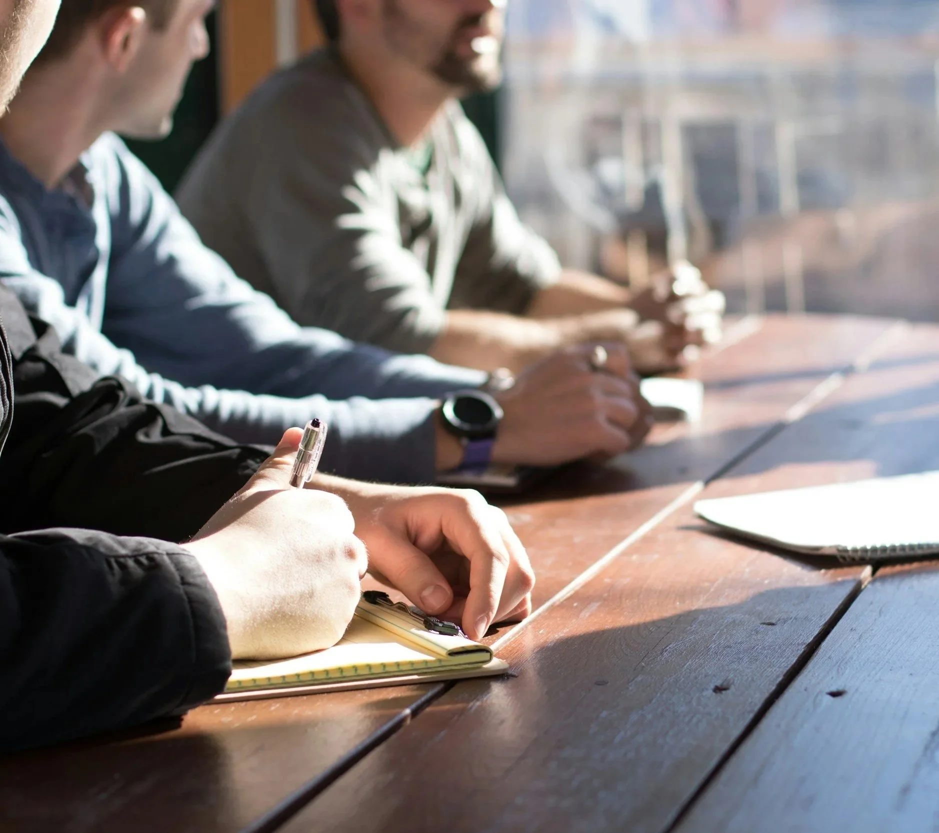 Close-up of three people sitting at a table, taking notes with pens and notebooks, with sunlight streaming through a window.