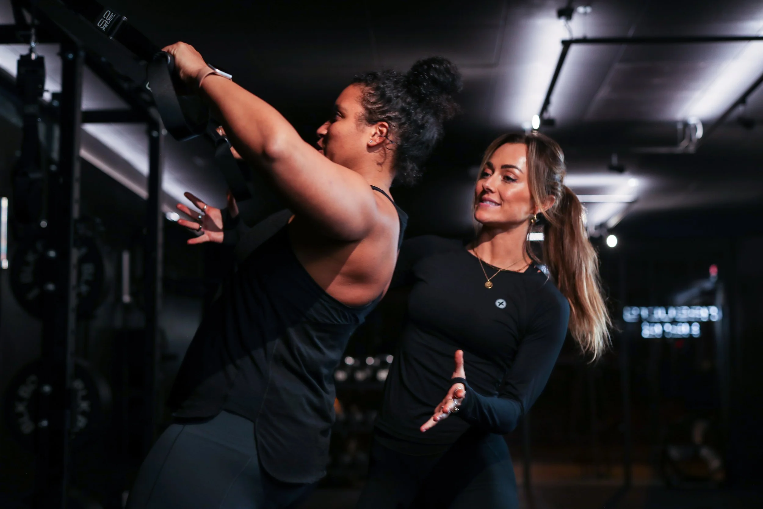 Two women in black workout clothes engaging in a fitness class or exercise session in a gym, focusing on resistance band exercises.