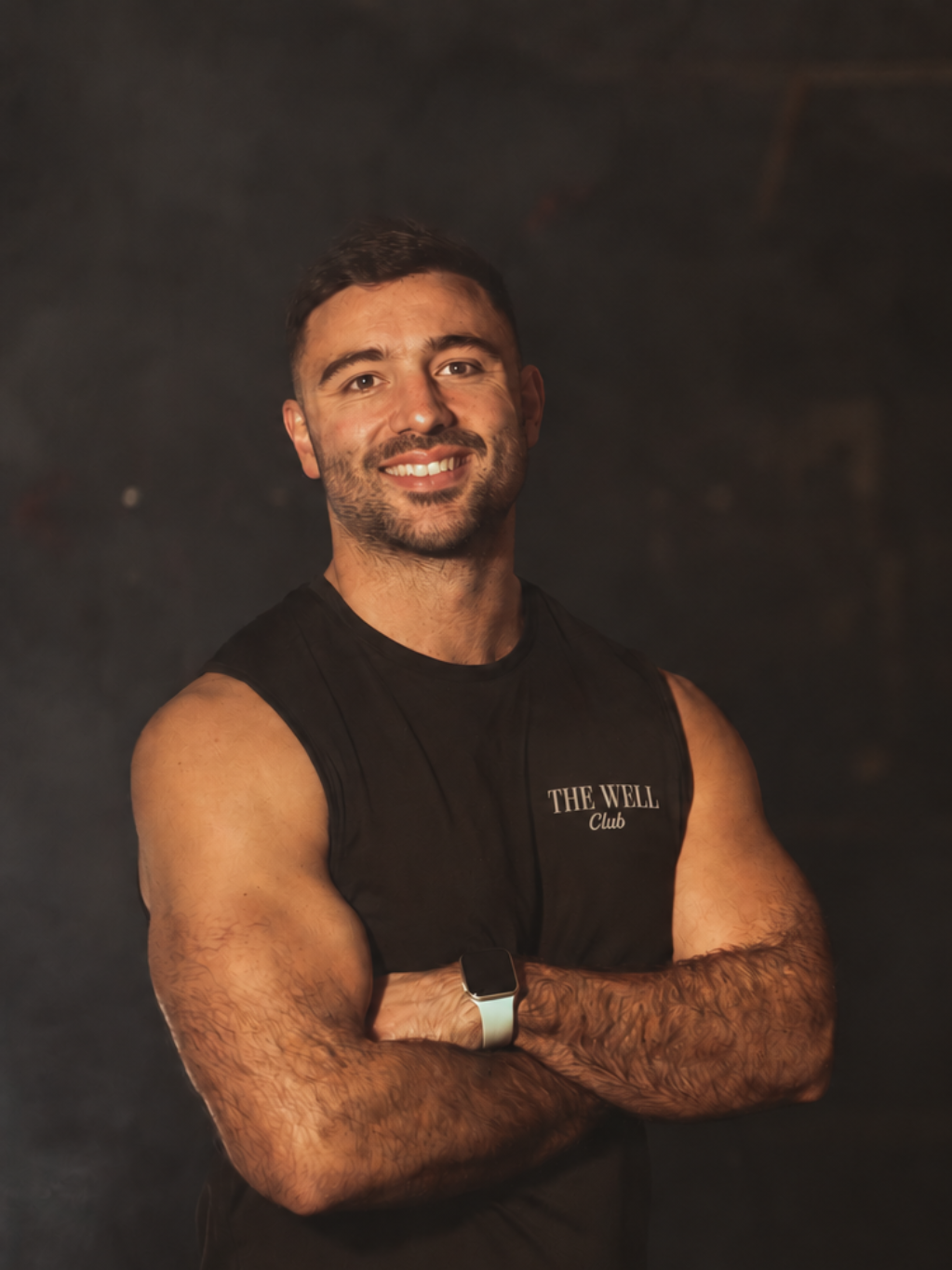 A smiling man with short dark hair and a beard, wearing a sleeveless black shirt with 'THE WELL Club' written on it, standing with arms crossed against a dark background.